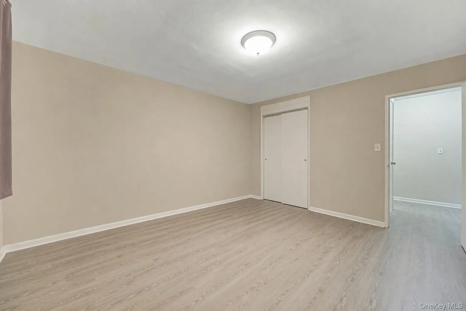Room featuring light wood-finish flooring, neutral wall paint, white baseboards, a ceiling-mounted light fixture, and a built-in closet with bi-fold doors Room featuring light wood-finish flooring, neutral wall paint, white baseboards, a ceiling-mounted light fixture, and a built-in closet with bi-fold doors