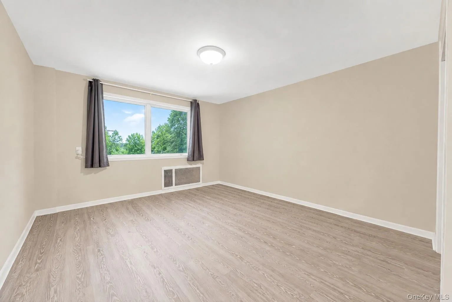 Spacious room with light-toned walls, wood-finish flooring, a large window, window-mounted air conditioning unit, and a flush-mount ceiling light Spacious room with light-toned walls, wood-finish flooring, a large window, window-mounted air conditioning unit, and a flush-mount ceiling light