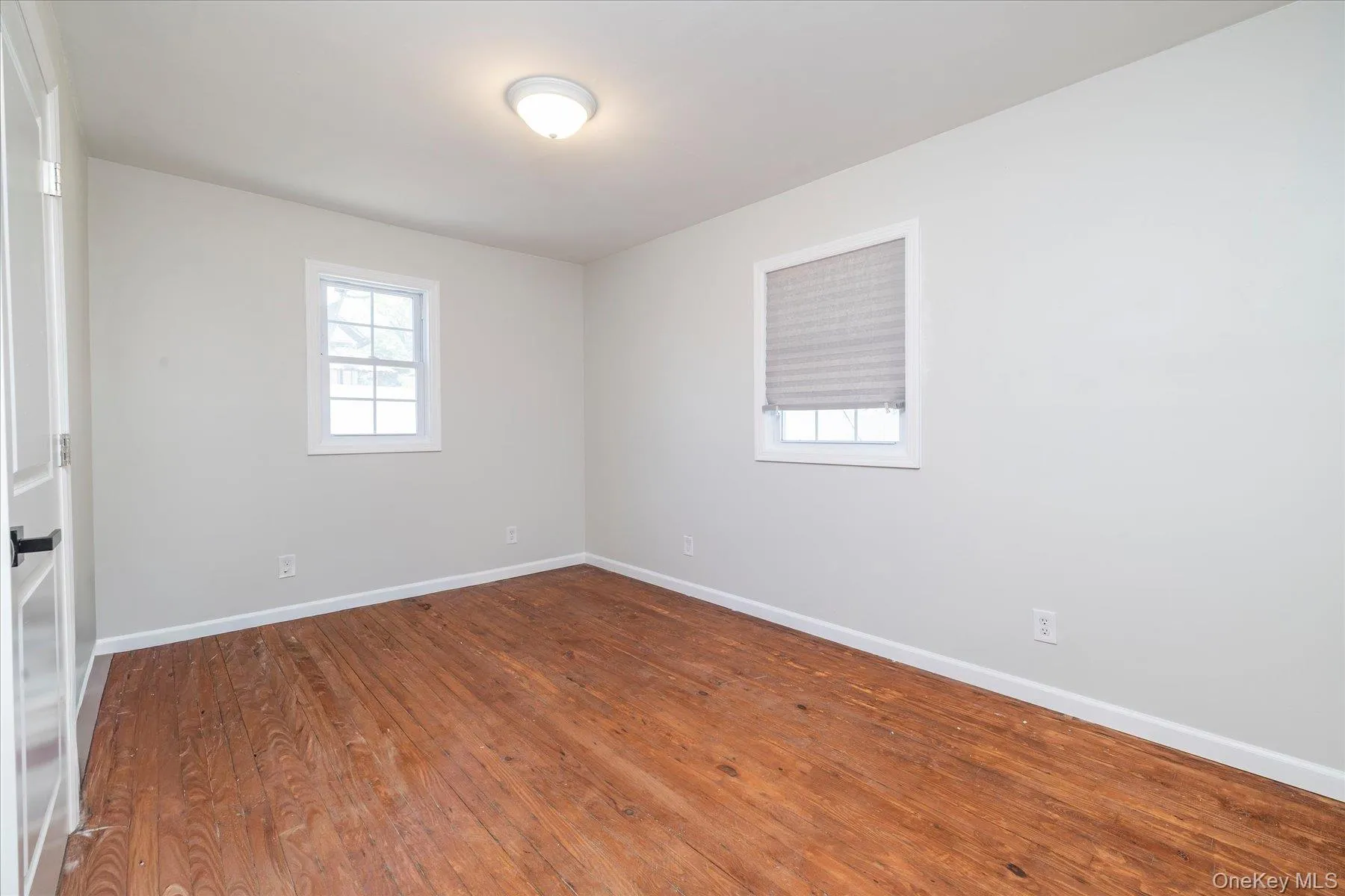 Bright room featuring wood-finish flooring, light-toned walls, two windows with white trim, and a flush-mount ceiling light fixture Bright room featuring wood-finish flooring, light-toned walls, two windows with white trim, and a flush-mount ceiling light fixture
