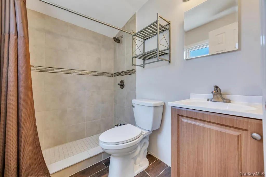 Bathroom featuring a tiled shower with decorative accent border, a white toilet, and a wood-finish vanity with an integrated sink and wall-mounted mirror Bathroom featuring a tiled shower with decorative accent border, a white toilet, and a wood-finish vanity with an integrated sink and wall-mounted mirror