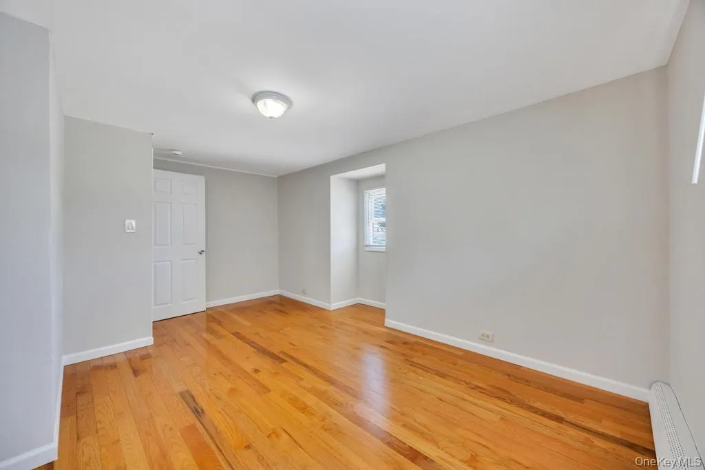Room featuring wood-finish flooring, light gray wall paint, a white paneled door, a single window, and a ceiling-mounted light fixture Room featuring wood-finish flooring, light gray wall paint, a white paneled door, a single window, and a ceiling-mounted light fixture