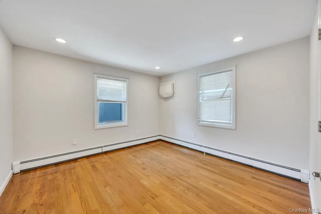 Room featuring hardwood flooring, recessed lighting, two windows with blinds, and white baseboard heating Room featuring hardwood flooring, recessed lighting, two windows with blinds, and white baseboard heating