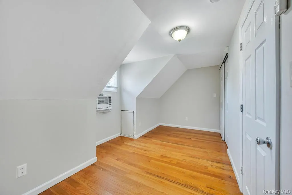 Attic space featuring wood-finish flooring, a window-mounted air conditioning unit, white trim, and a flush-mount ceiling light fixture Attic space featuring wood-finish flooring, a window-mounted air conditioning unit, white trim, and a flush-mount ceiling light fixture