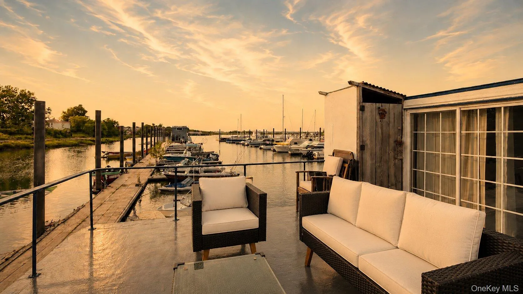 Outdoor deck with water access, featuring sliding glass doors, a weathered wood-paneled utility structure, and a black metal railing overlooking a marina with boats Outdoor deck with water access, featuring sliding glass doors, a weathered wood-paneled utility structure, and a black metal railing overlooking a marina with boats
