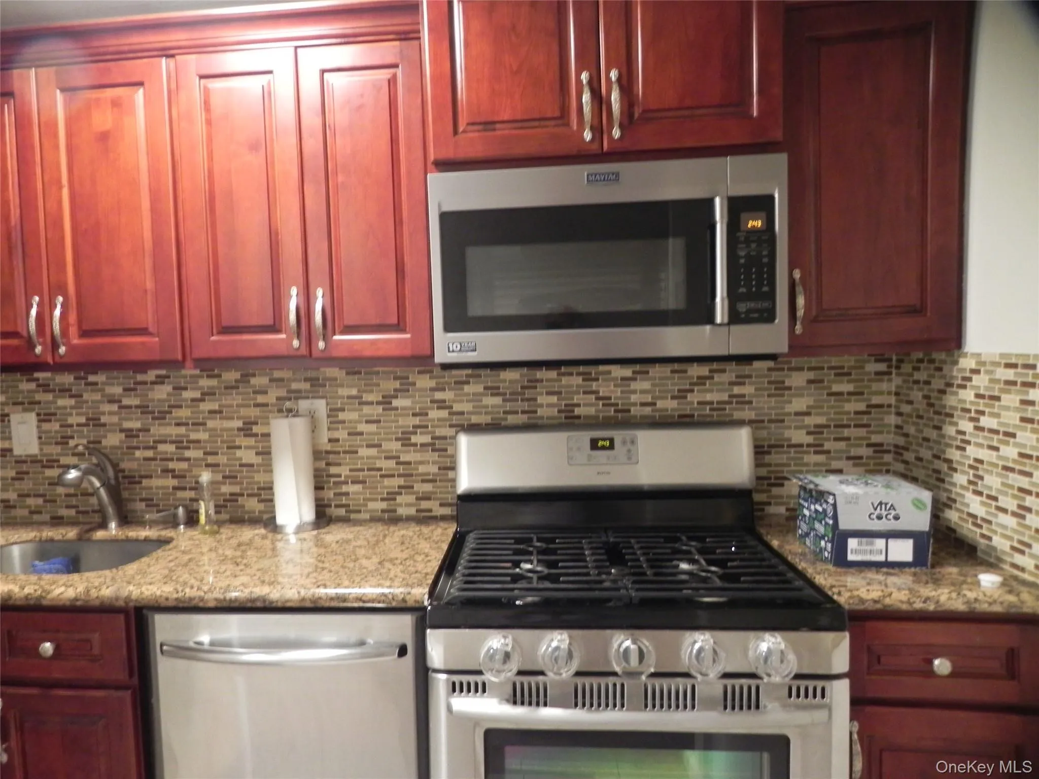 Kitchen featuring stainless steel appliances, reddish brown cabinets, backsplash, and light stone countertops Kitchen featuring stainless steel appliances, reddish brown cabinets, backsplash, and light stone countertops