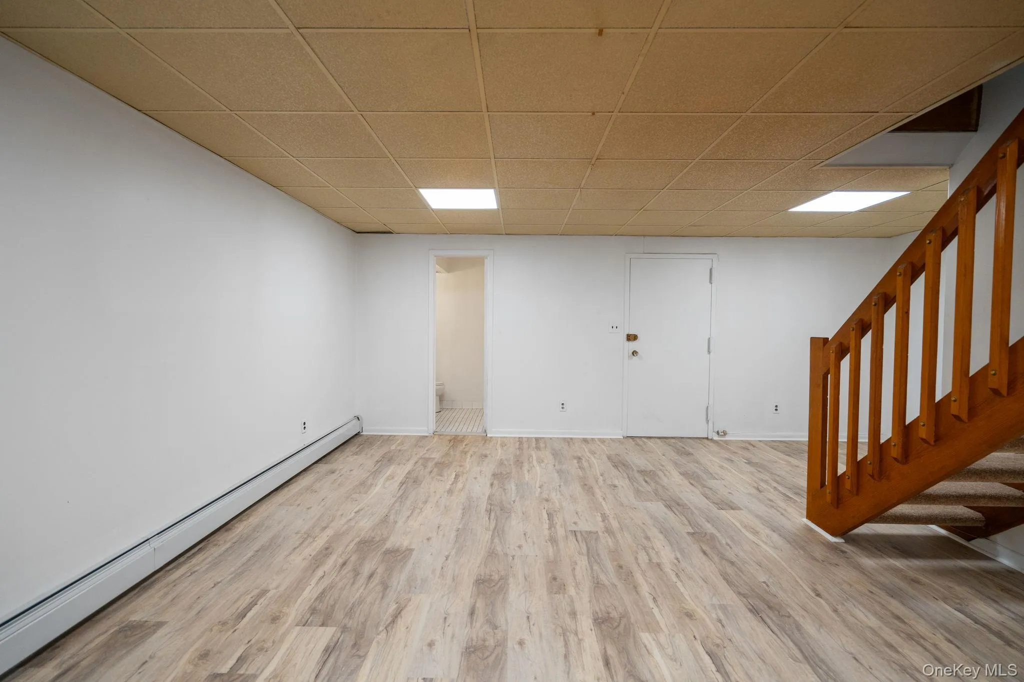 Spacious finished basement featuring wood-finish flooring, white walls, and a light-colored acoustical tile ceiling Spacious finished basement featuring wood-finish flooring, white walls, and a light-colored acoustical tile ceiling