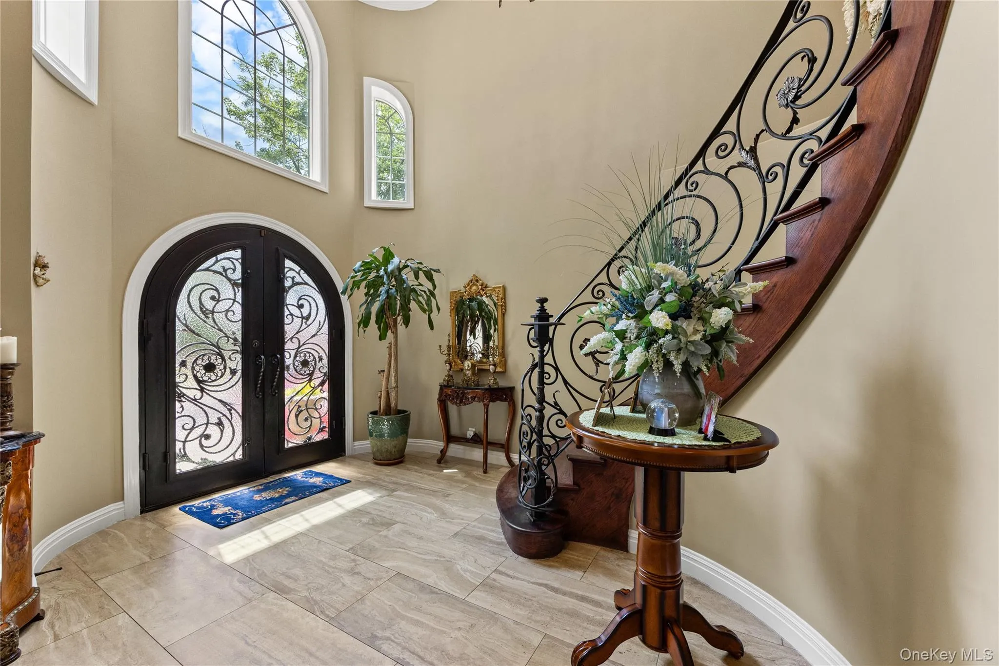 Grand foyer featuring arched double entry doors with intricate wrought iron detailing, large arched windows, a curved staircase with wrought iron railing, and stone-look tile flooring Grand foyer featuring arched double entry doors with intricate wrought iron detailing, large arched windows, a curved staircase with wrought iron railing, and stone-look tile flooring