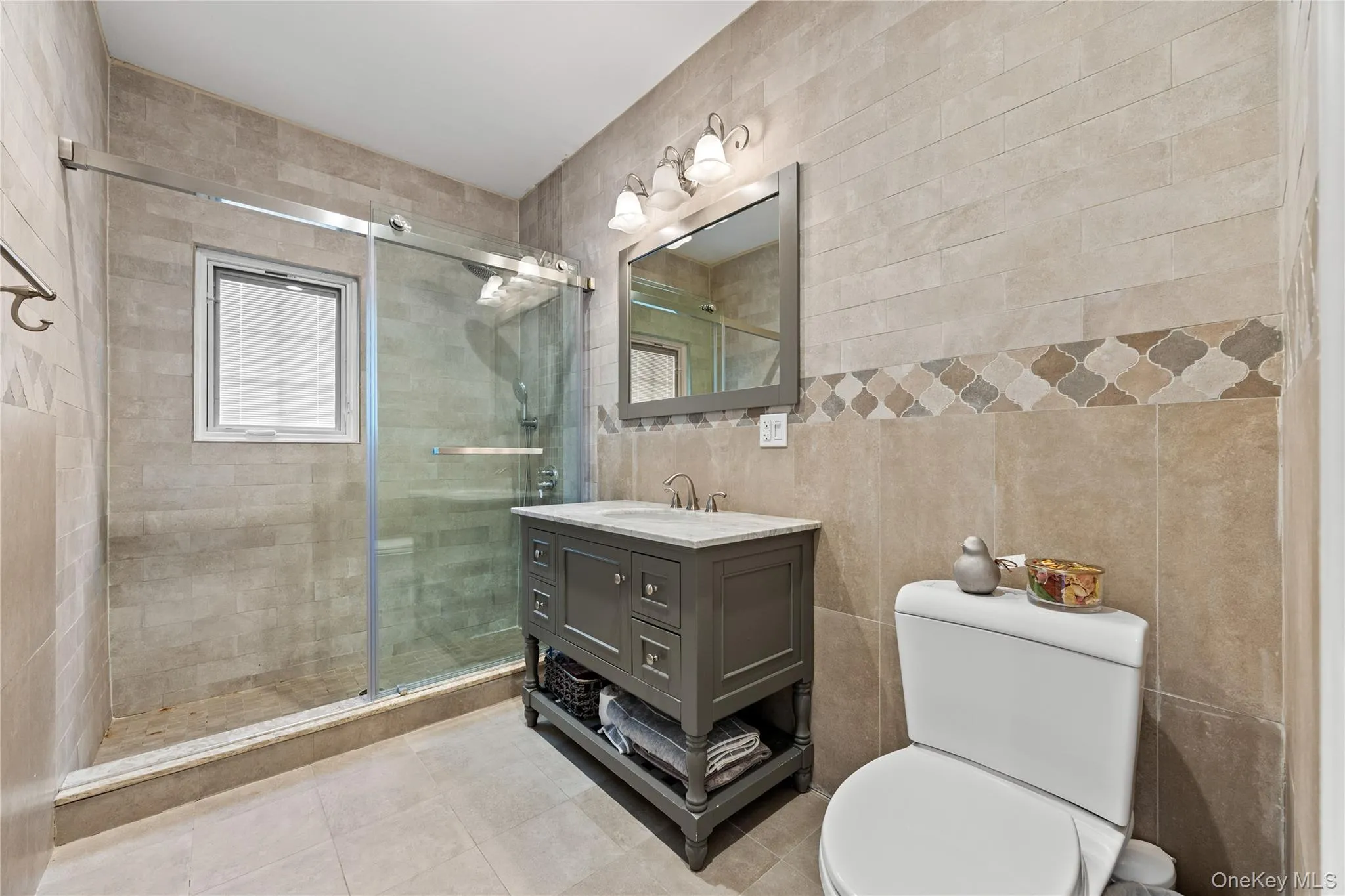 Tiled bathroom featuring a glass-enclosed shower with a window, a vanity with storage, and a light-colored tile floor Tiled bathroom featuring a glass-enclosed shower with a window, a vanity with storage, and a light-colored tile floor