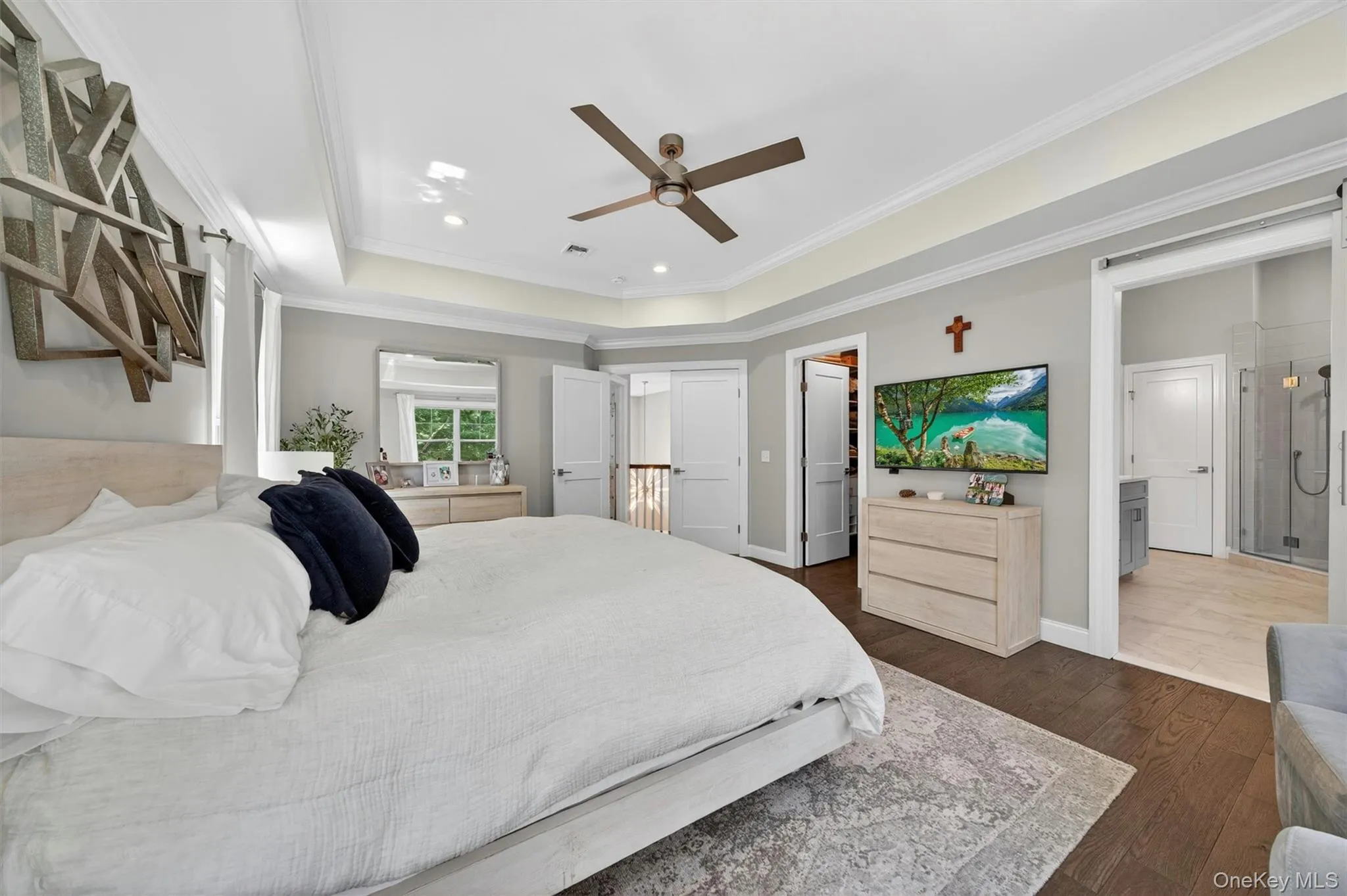 Hardwood flooring throughout, tray ceiling with recessed lighting and a ceiling fan, dual white doors opening to a hallway, a sliding barn door entryway to a bathroom with a glass-enclosed shower, and a window with outdoor greenery visible Hardwood flooring throughout, tray ceiling with recessed lighting and a ceiling fan, dual white doors opening to a hallway, a sliding barn door entryway to a bathroom with a glass-enclosed shower, and a window with outdoor greenery visible