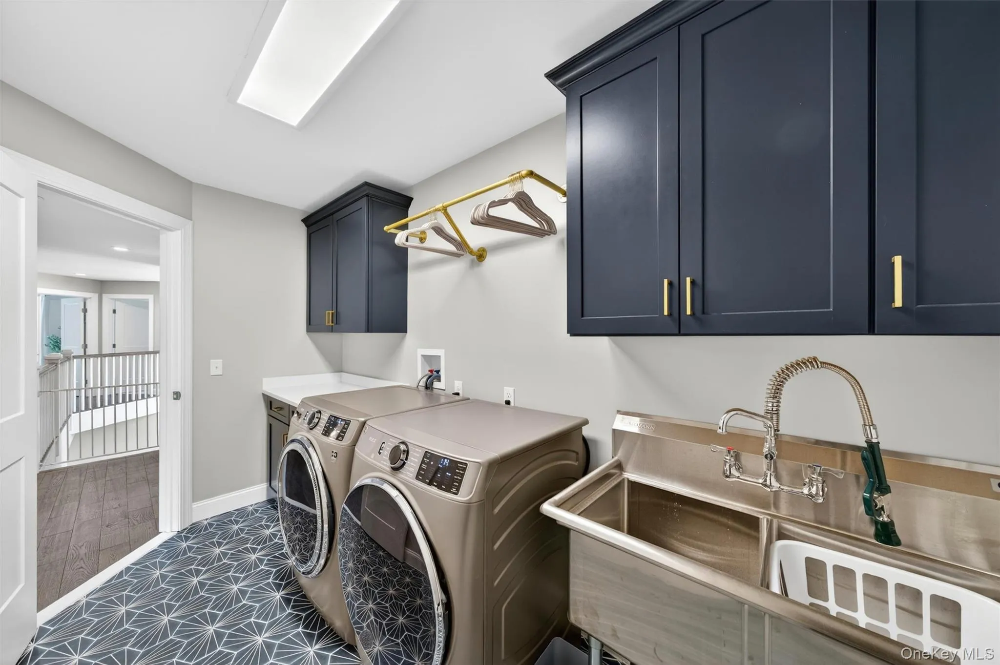 Laundry room featuring bold patterned floor tile, dark shaker-style cabinetry with gold-toned hardware, a stainless steel utility sink with a pull-down faucet, a gold-toned hanging rod, and recessed lighting Laundry room featuring bold patterned floor tile, dark shaker-style cabinetry with gold-toned hardware, a stainless steel utility sink with a pull-down faucet, a gold-toned hanging rod, and recessed lighting