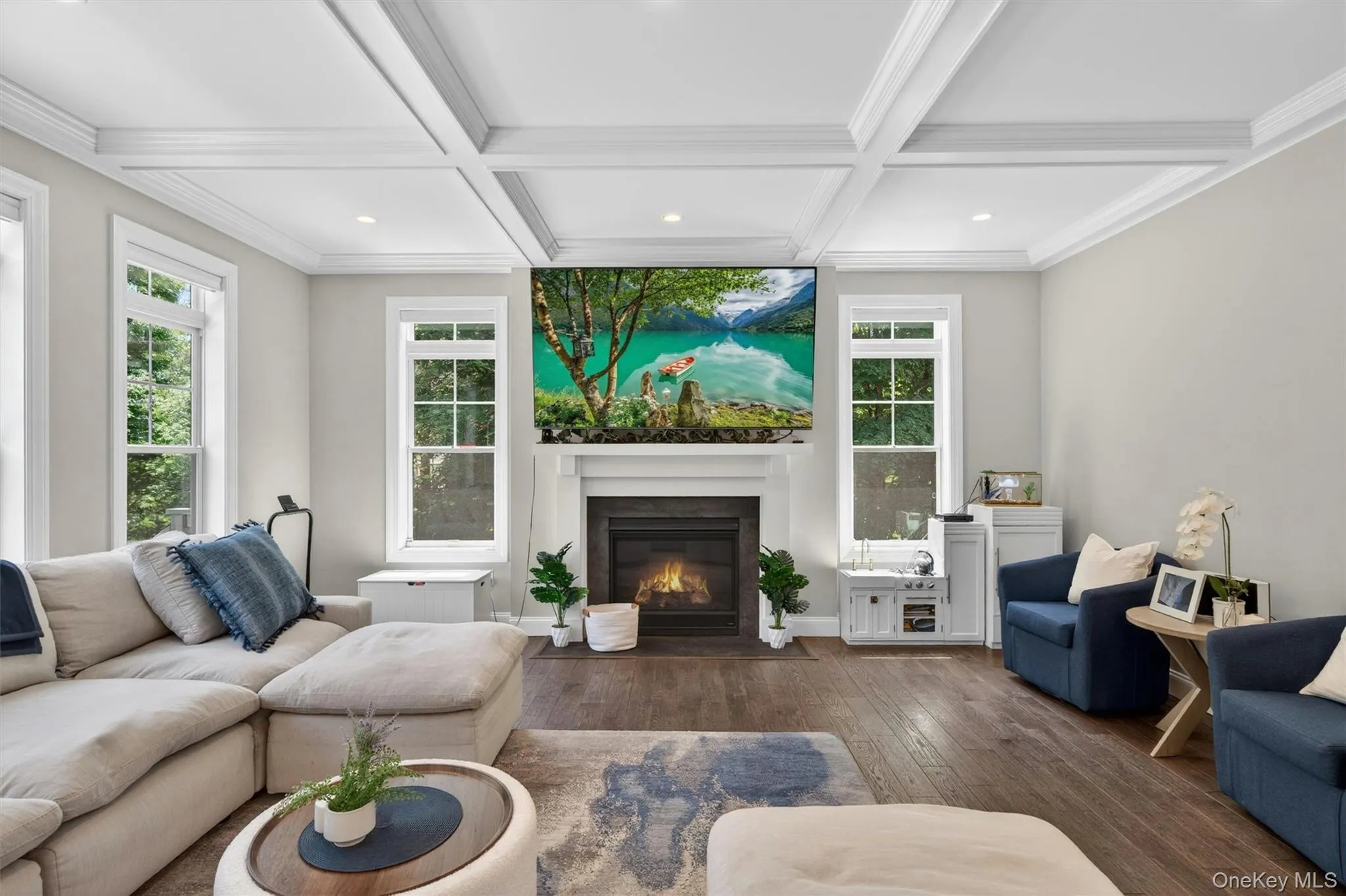 Spacious living area featuring a fireplace with a white mantel, coffered ceilings, dark wood-finish flooring, recessed lighting, and multiple windows Spacious living area featuring a fireplace with a white mantel, coffered ceilings, dark wood-finish flooring, recessed lighting, and multiple windows