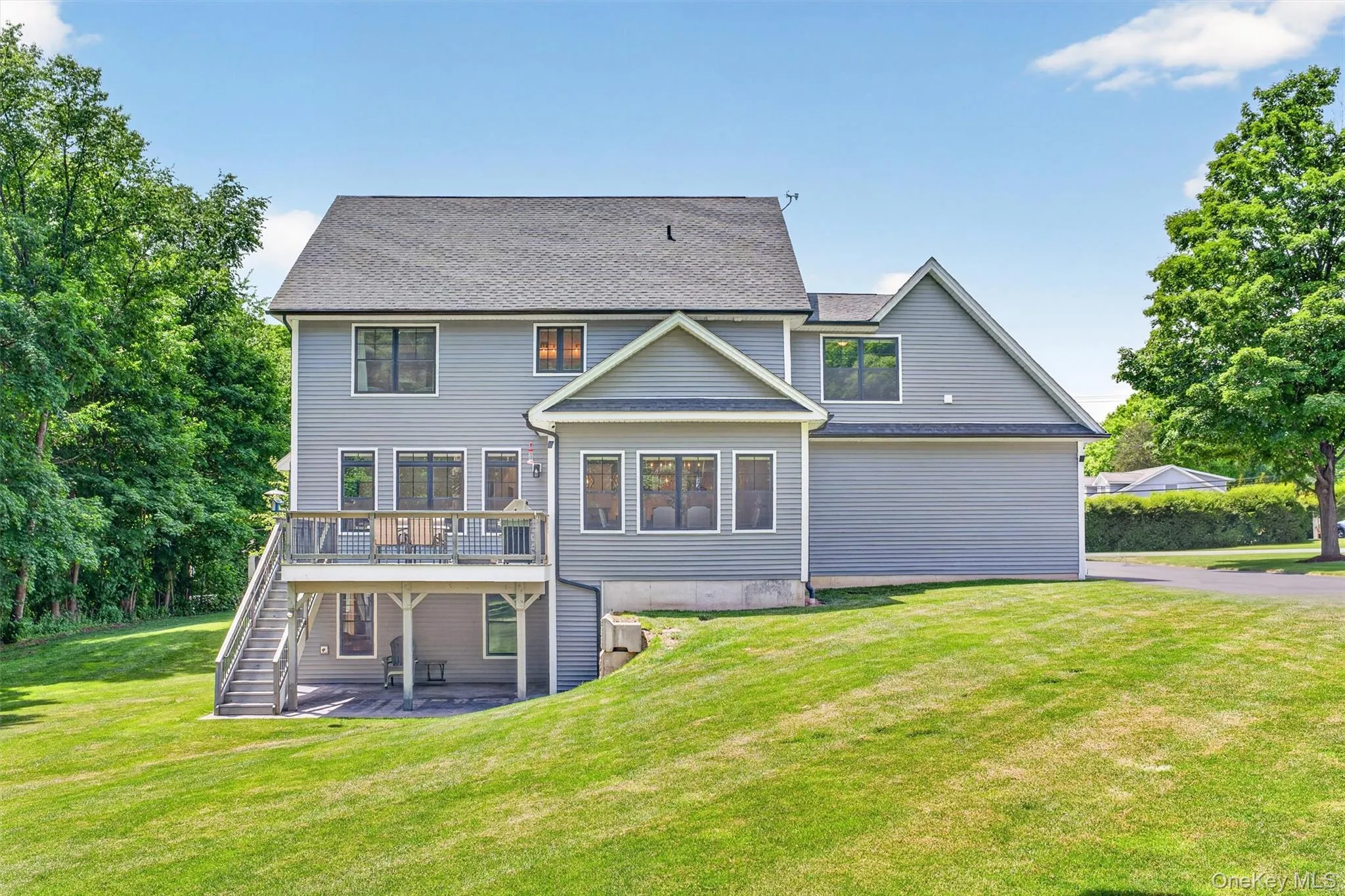 Rear exterior featuring gray siding, a composite deck with railing, stairs to a lower patio, and a sloped lawn Rear exterior featuring gray siding, a composite deck with railing, stairs to a lower patio, and a sloped lawn