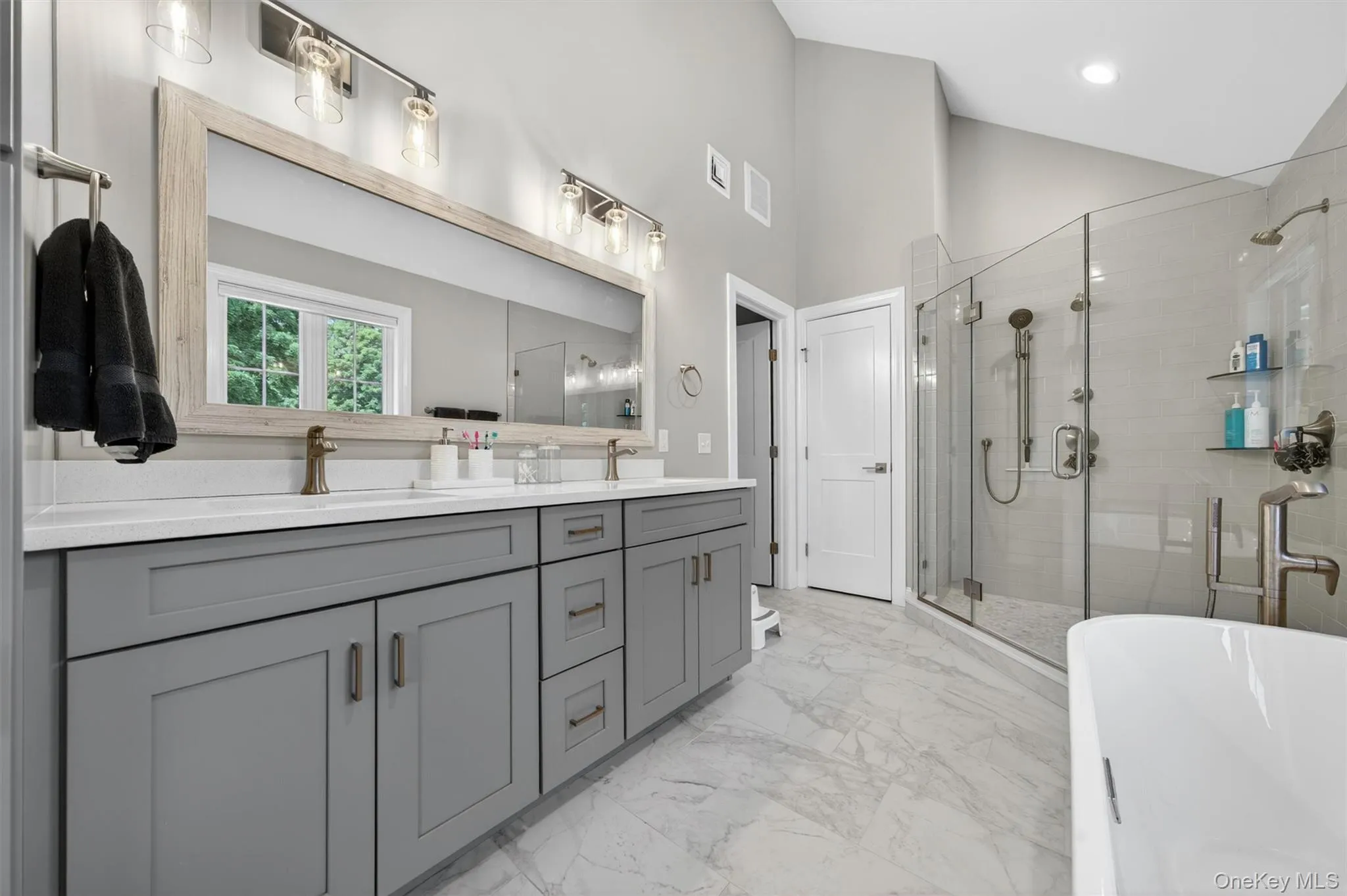 Light-filled bathroom featuring a dual vanity with gray cabinetry and white countertops, a large framed mirror, a spacious glass-enclosed shower, and a freestanding soaking tub Light-filled bathroom featuring a dual vanity with gray cabinetry and white countertops, a large framed mirror, a spacious glass-enclosed shower, and a freestanding soaking tub