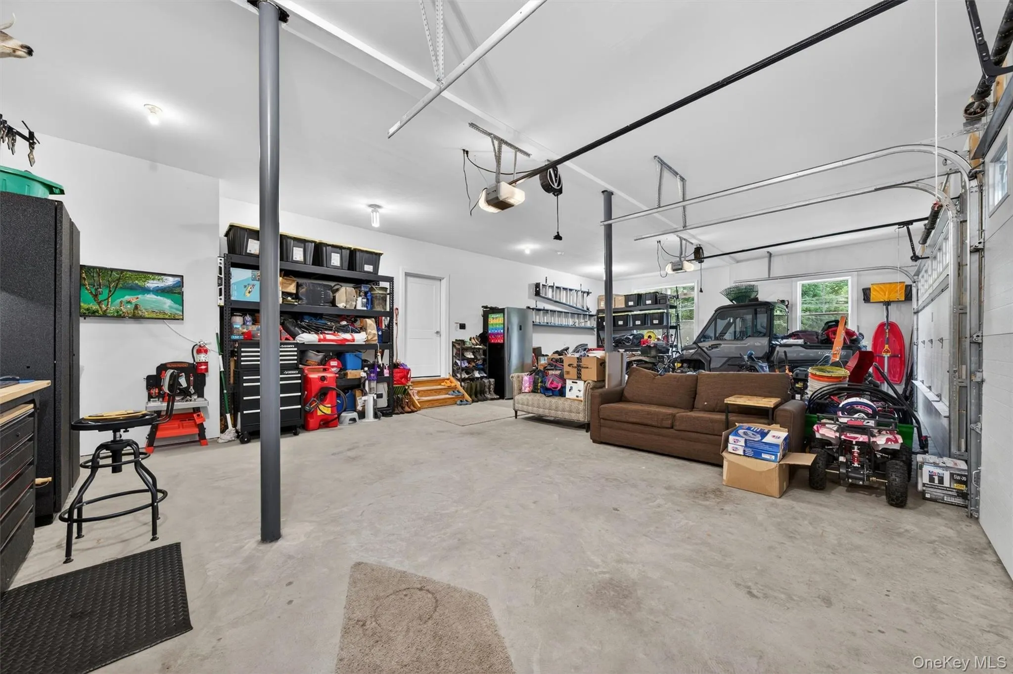 Spacious garage featuring concrete flooring, white walls, multiple overhead lighting fixtures, and a garage door with track system Spacious garage featuring concrete flooring, white walls, multiple overhead lighting fixtures, and a garage door with track system