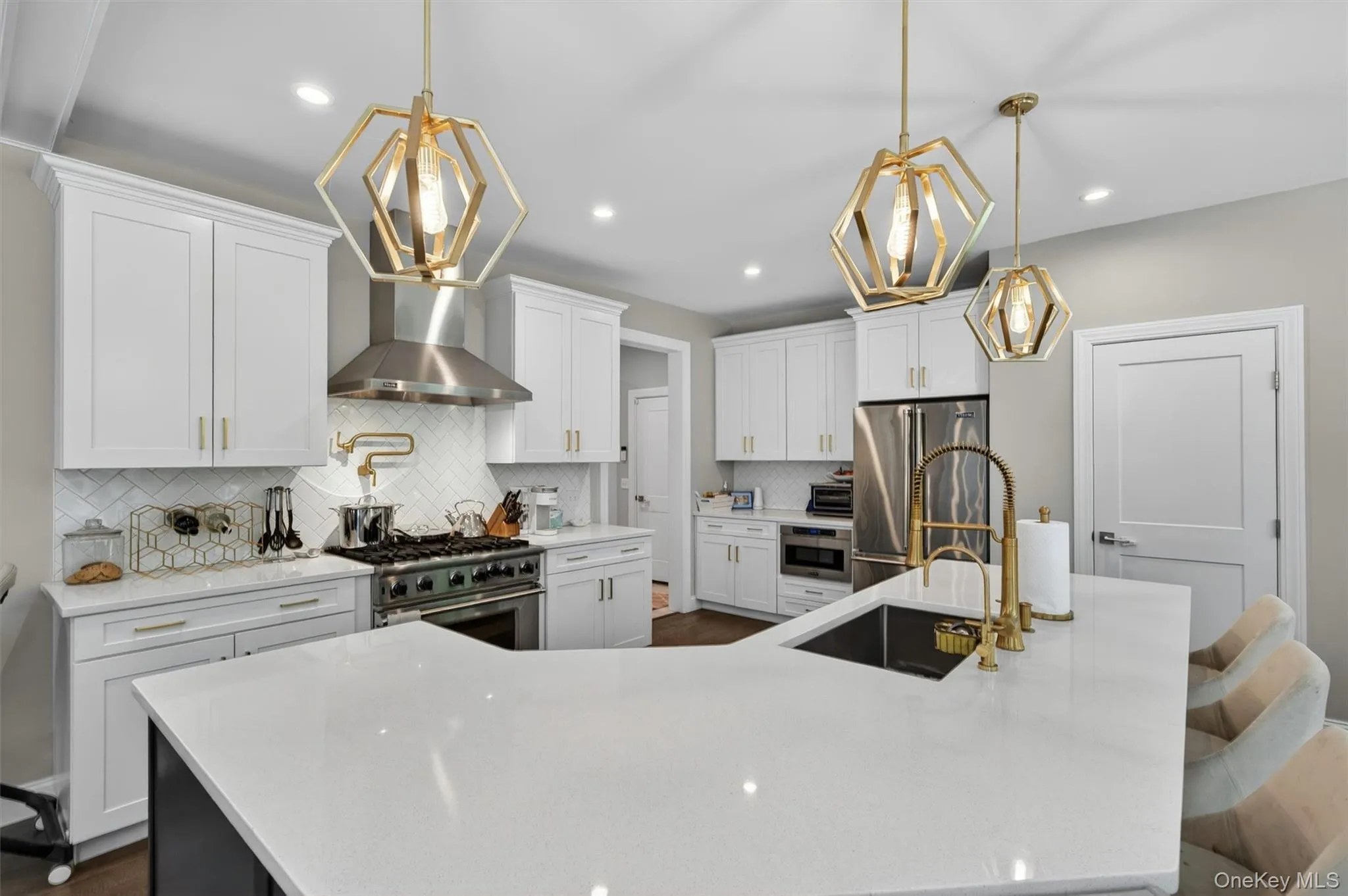 Kitchen featuring a large central island with a sink and gold-tone faucet, white shaker-style cabinetry, stainless steel appliances, a herringbone tile backsplash, and decorative pendant lighting Kitchen featuring a large central island with a sink and gold-tone faucet, white shaker-style cabinetry, stainless steel appliances, a herringbone tile backsplash, and decorative pendant lighting