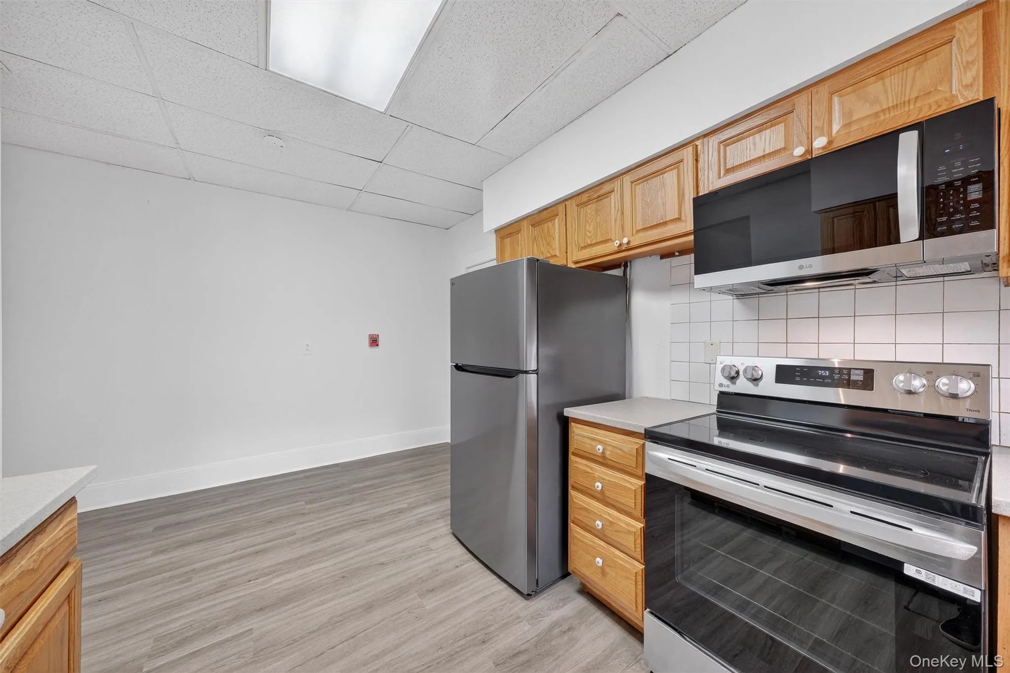Kitchen featuring stainless steel appliances, a drop ceiling, light wood-style floors, decorative backsplash, and light countertops Kitchen featuring stainless steel appliances, a drop ceiling, light wood-style floors, decorative backsplash, and light countertops