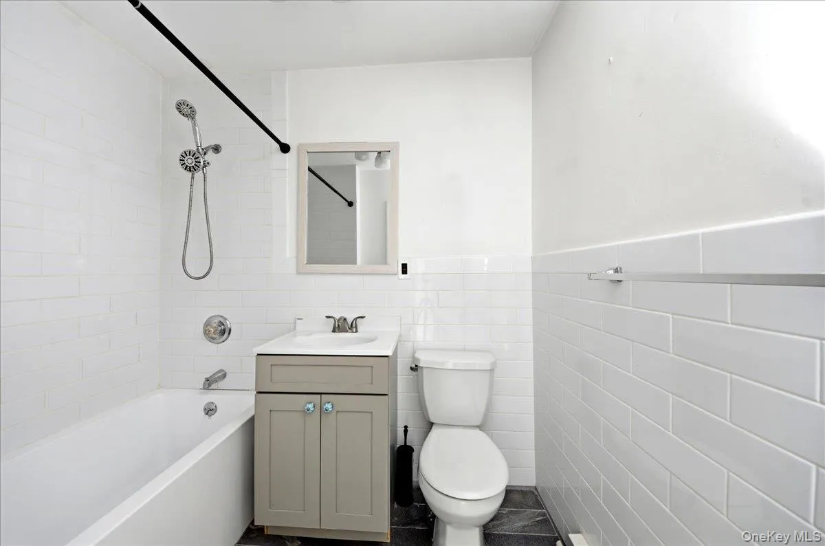 Bathroom featuring white subway tile walls, a bathtub with an integrated showerhead, a vanity with a white countertop and gray cabinetry, a wall-mounted mirror, and dark tile flooring Bathroom featuring white subway tile walls, a bathtub with an integrated showerhead, a vanity with a white countertop and gray cabinetry, a wall-mounted mirror, and dark tile flooring