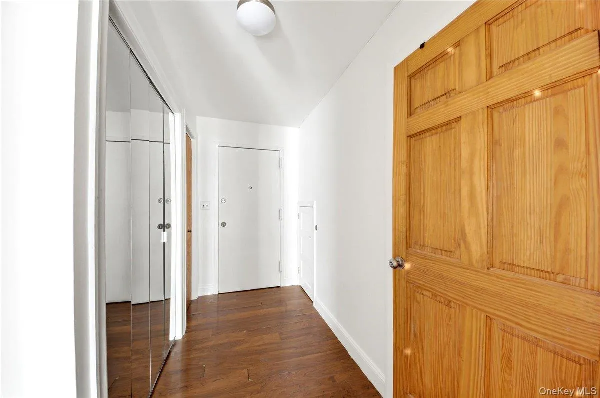 Hallway featuring wood-finish flooring, a paneled wooden door, mirrored closet doors, and white walls Hallway featuring wood-finish flooring, a paneled wooden door, mirrored closet doors, and white walls