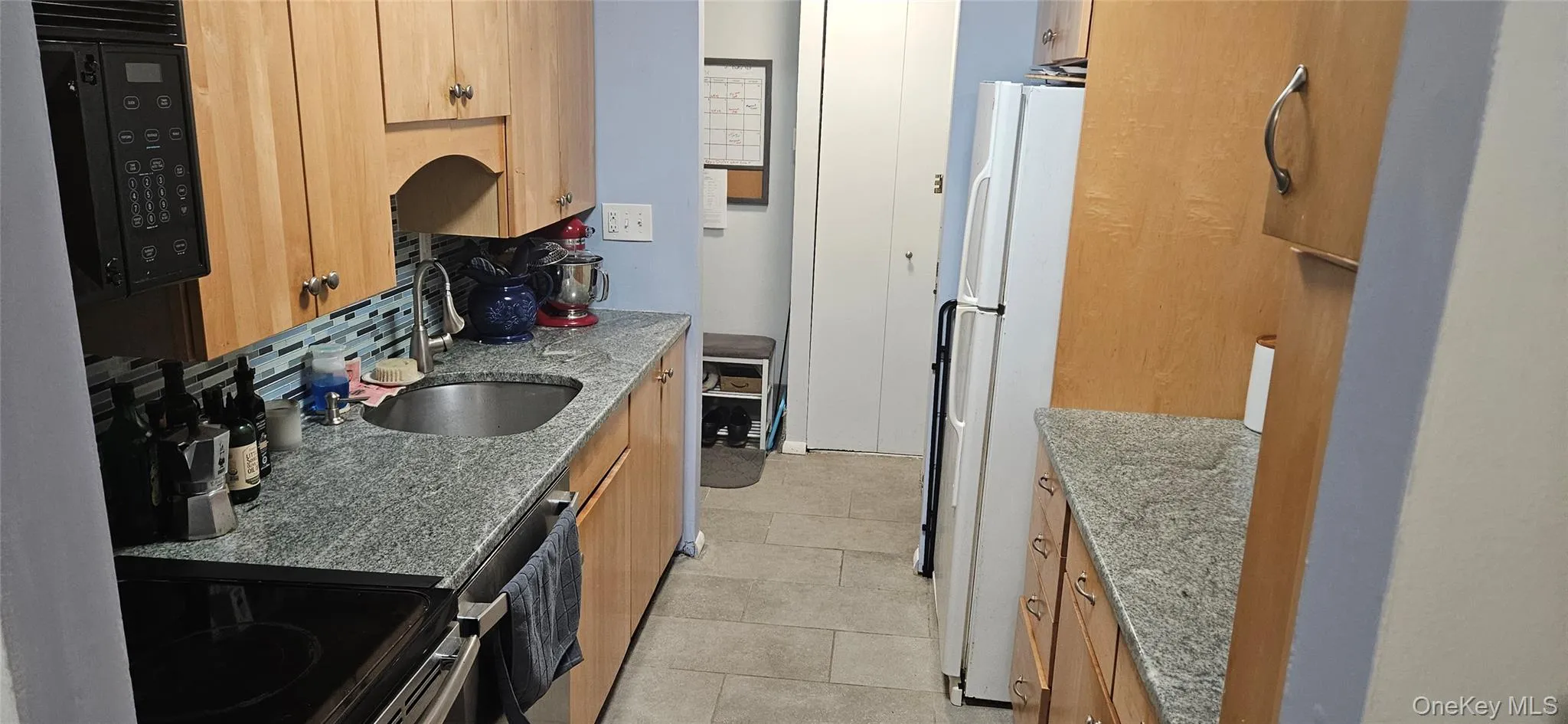 Galley kitchen featuring light wood-finish cabinetry, speckled countertops, a mosaic tile backsplash, and tile flooring Galley kitchen featuring light wood-finish cabinetry, speckled countertops, a mosaic tile backsplash, and tile flooring