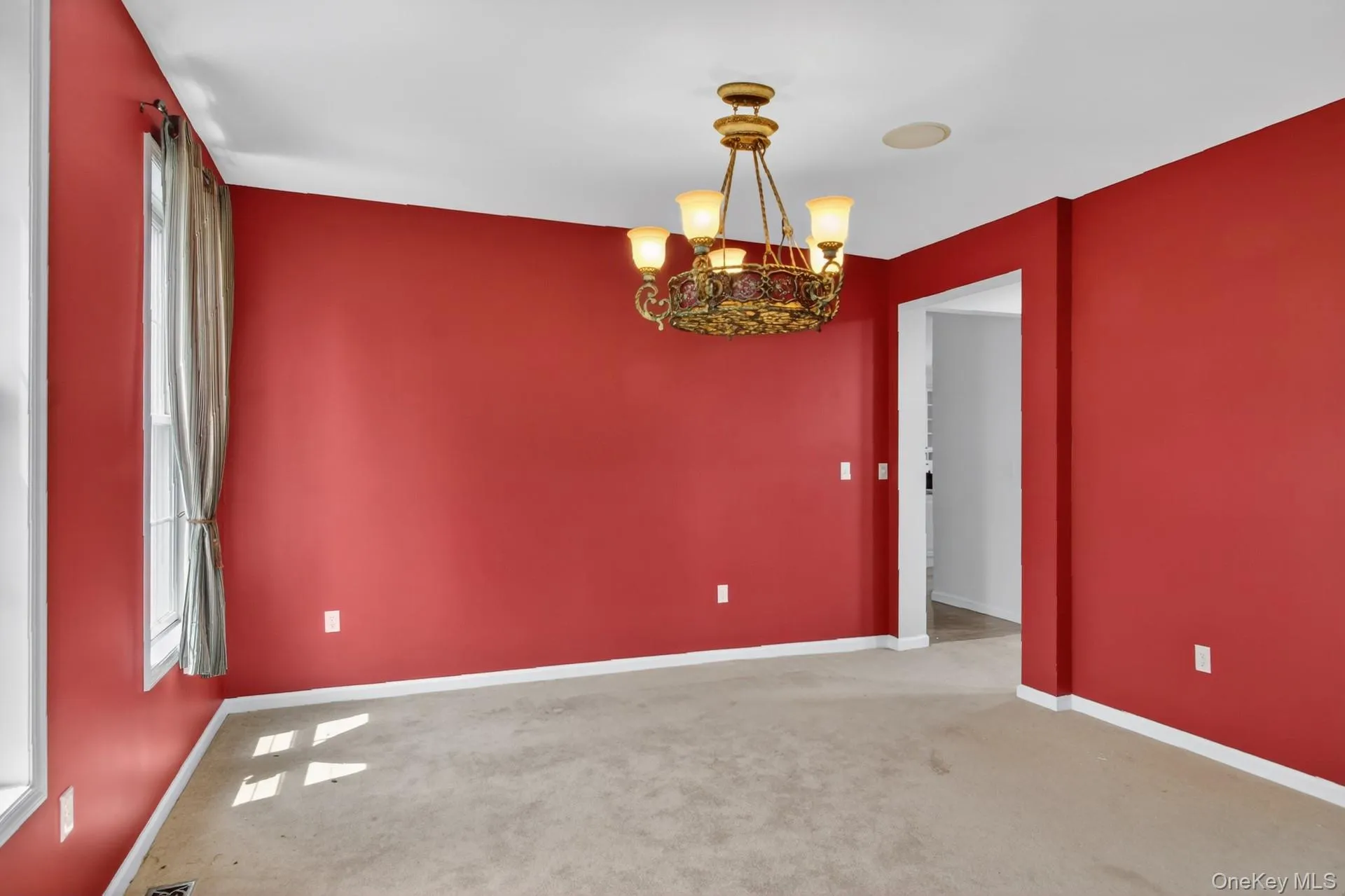 Spacious interior room featuring red walls, light beige carpeting, and a decorative brass chandelier Spacious interior room featuring red walls, light beige carpeting, and a decorative brass chandelier