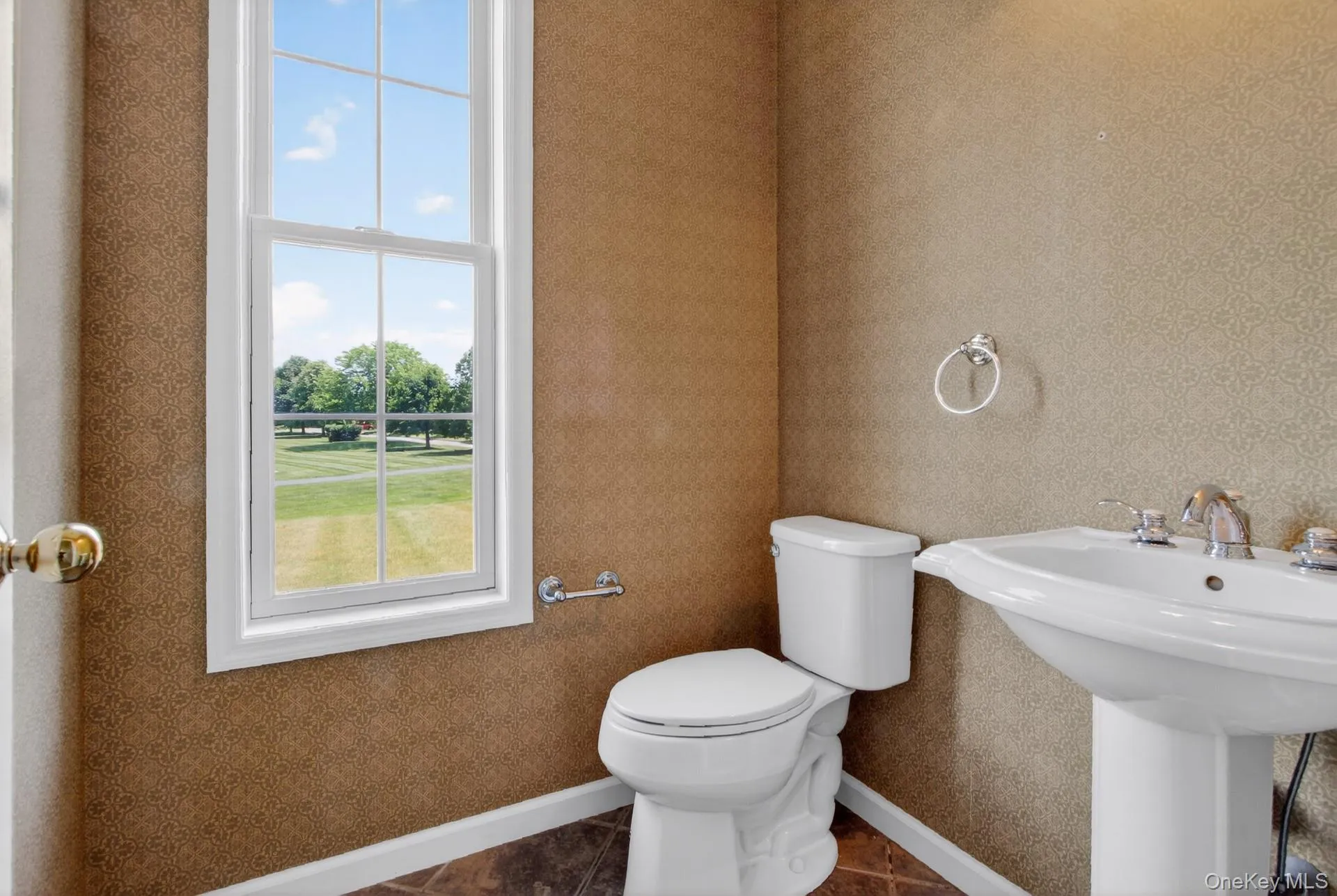Pedestal sink with chrome fixtures, white commode, patterned wall covering, wood-finish flooring, and a single-hung window with a verdant outdoor landscape Pedestal sink with chrome fixtures, white commode, patterned wall covering, wood-finish flooring, and a single-hung window with a verdant outdoor landscape