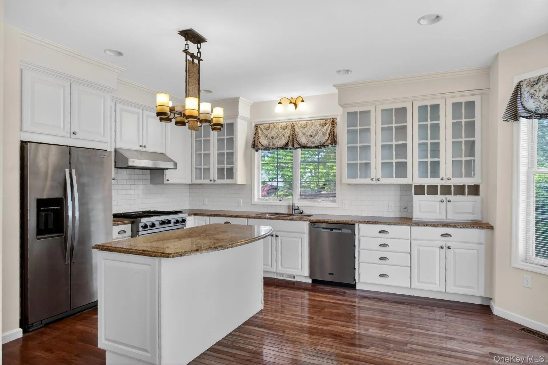 Kitchen featuring an island with a stone countertop, white cabinetry with glass-front uppers, stainless steel appliances, and wood-finish flooring Kitchen featuring an island with a stone countertop, white cabinetry with glass-front uppers, stainless steel appliances, and wood-finish flooring