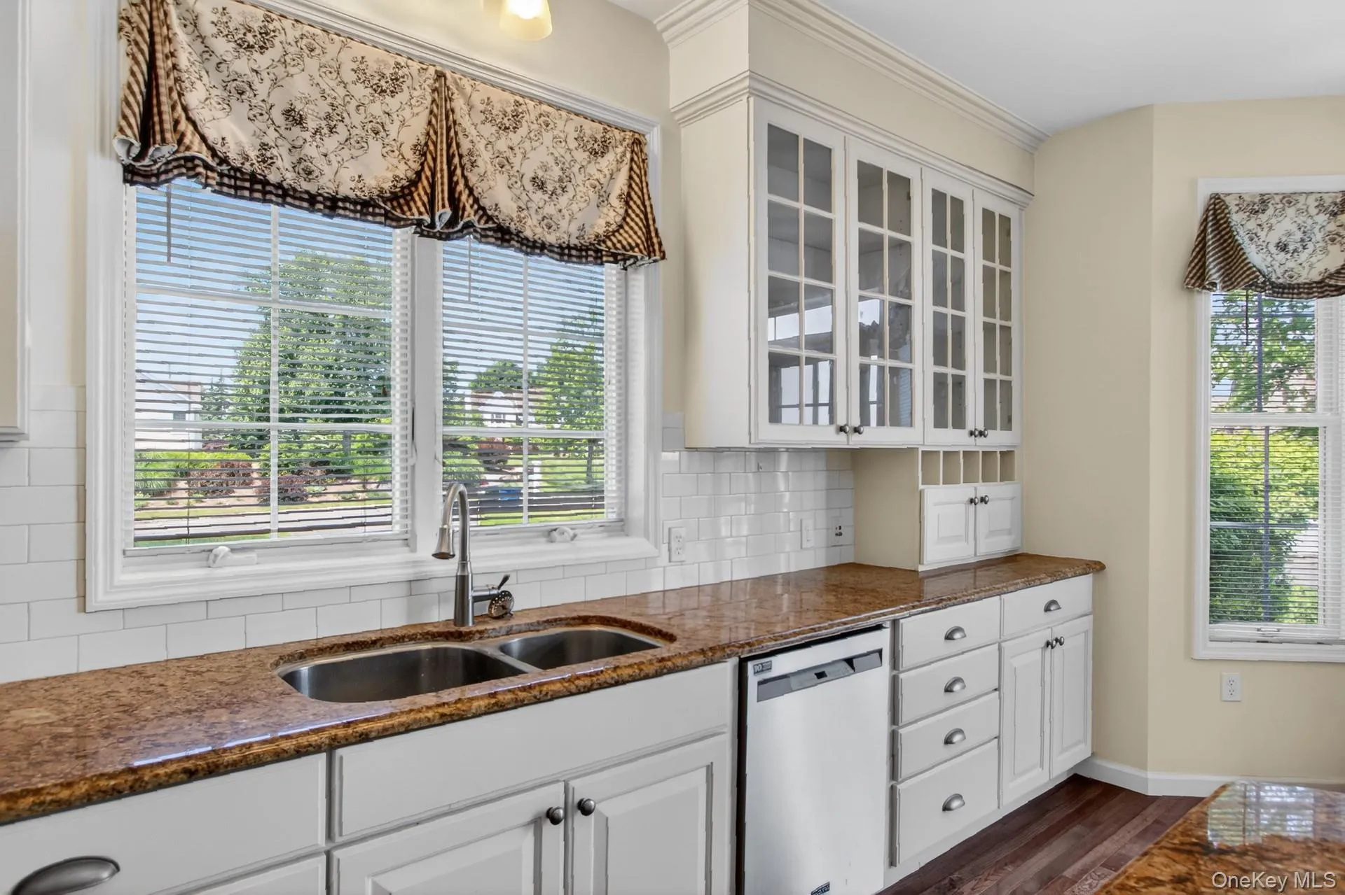 Kitchen featuring white cabinetry, brown granite-style countertops, stainless steel double basin sink, and white subway tile backsplash Kitchen featuring white cabinetry, brown granite-style countertops, stainless steel double basin sink, and white subway tile backsplash