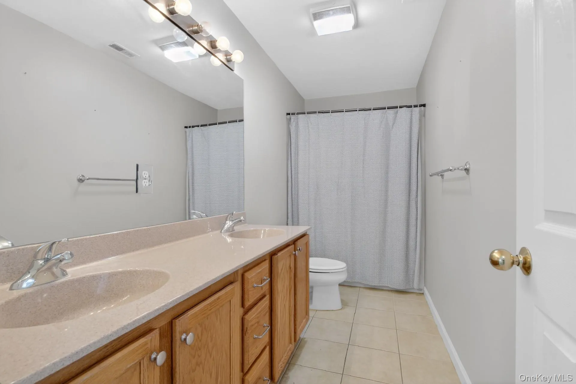 Bathroom featuring a double vanity with light wood cabinetry and integrated sinks, a large wall-mounted mirror, a tub/shower combination with a shower curtain, and neutral tile flooring Bathroom featuring a double vanity with light wood cabinetry and integrated sinks, a large wall-mounted mirror, a tub/shower combination with a shower curtain, and neutral tile flooring
