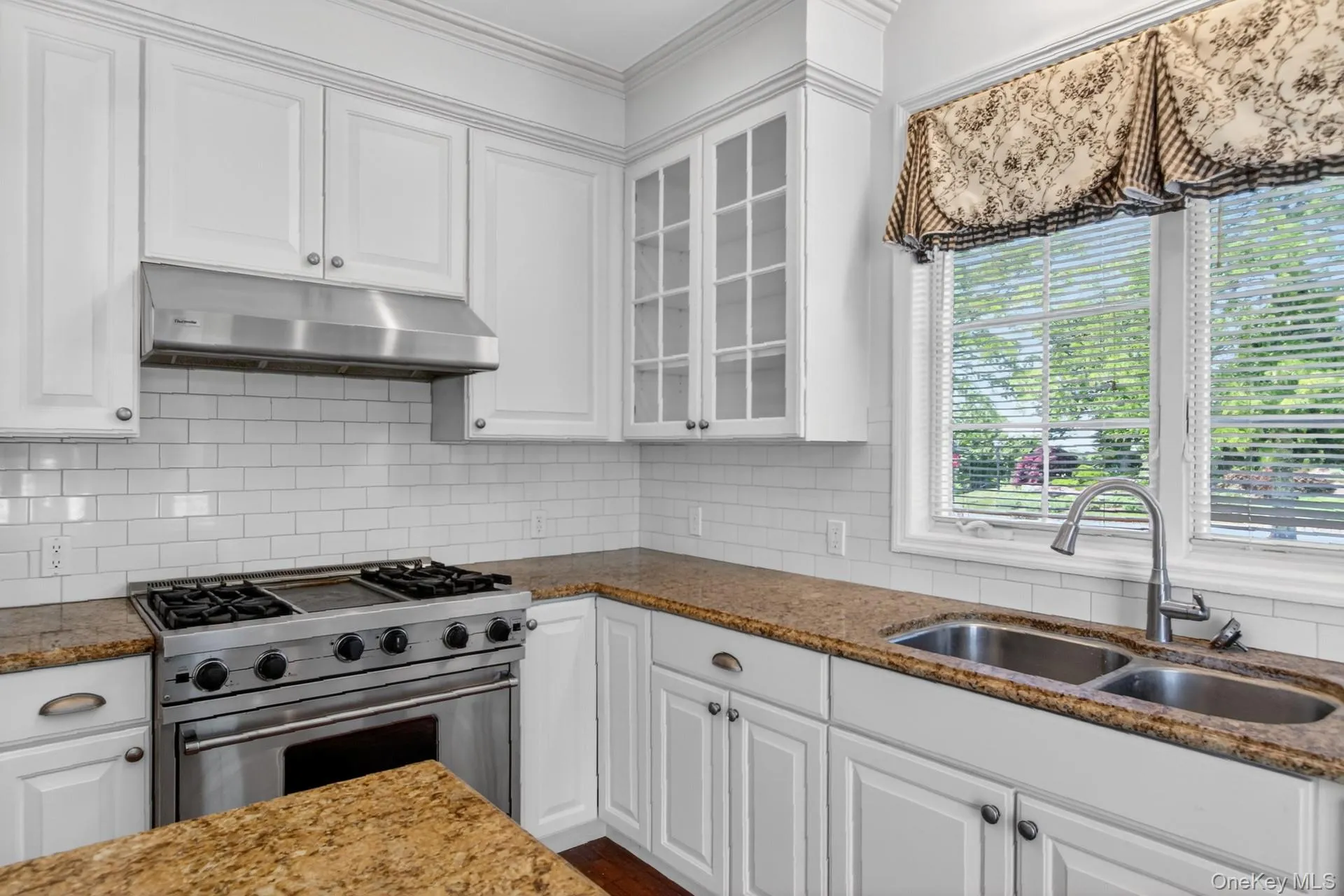 Kitchen featuring white cabinetry, subway tile backsplash, stainless steel range with hood, double basin sink with gooseneck faucet, and stone countertops Kitchen featuring white cabinetry, subway tile backsplash, stainless steel range with hood, double basin sink with gooseneck faucet, and stone countertops