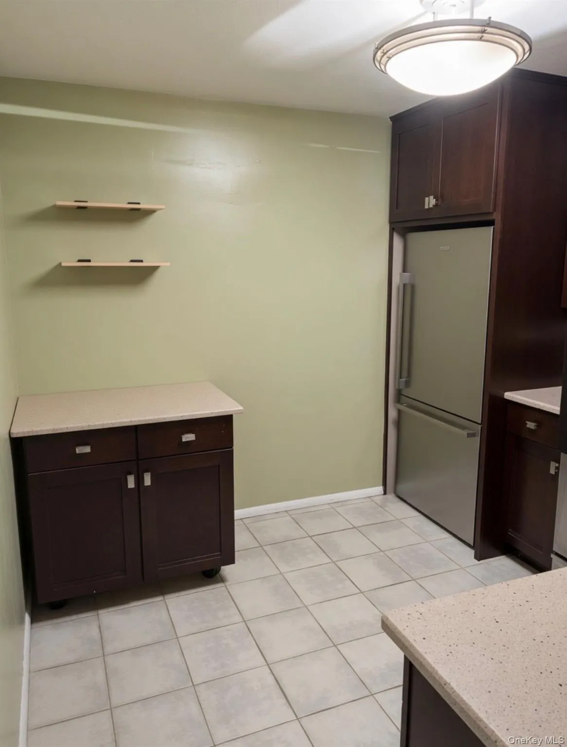 Kitchen area featuring tile flooring, dark wood-finish cabinetry, light-toned countertops, and a stainless steel refrigerator Kitchen area featuring tile flooring, dark wood-finish cabinetry, light-toned countertops, and a stainless steel refrigerator