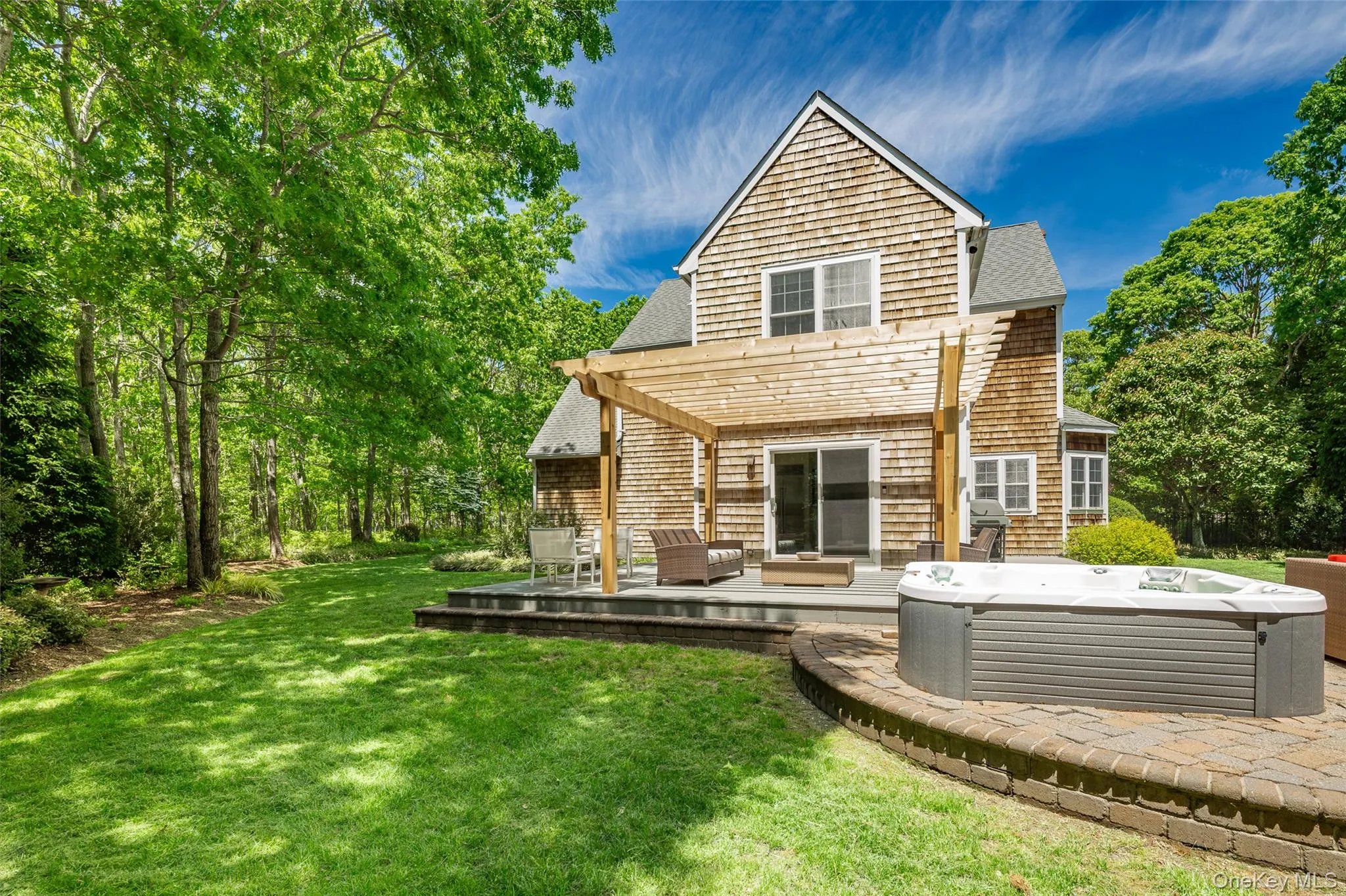 Spacious backyard featuring a hot tub on a paver patio with a stone retaining wall, a wood pergola over a patio area, and a cedar shingle exterior Spacious backyard featuring a hot tub on a paver patio with a stone retaining wall, a wood pergola over a patio area, and a cedar shingle exterior