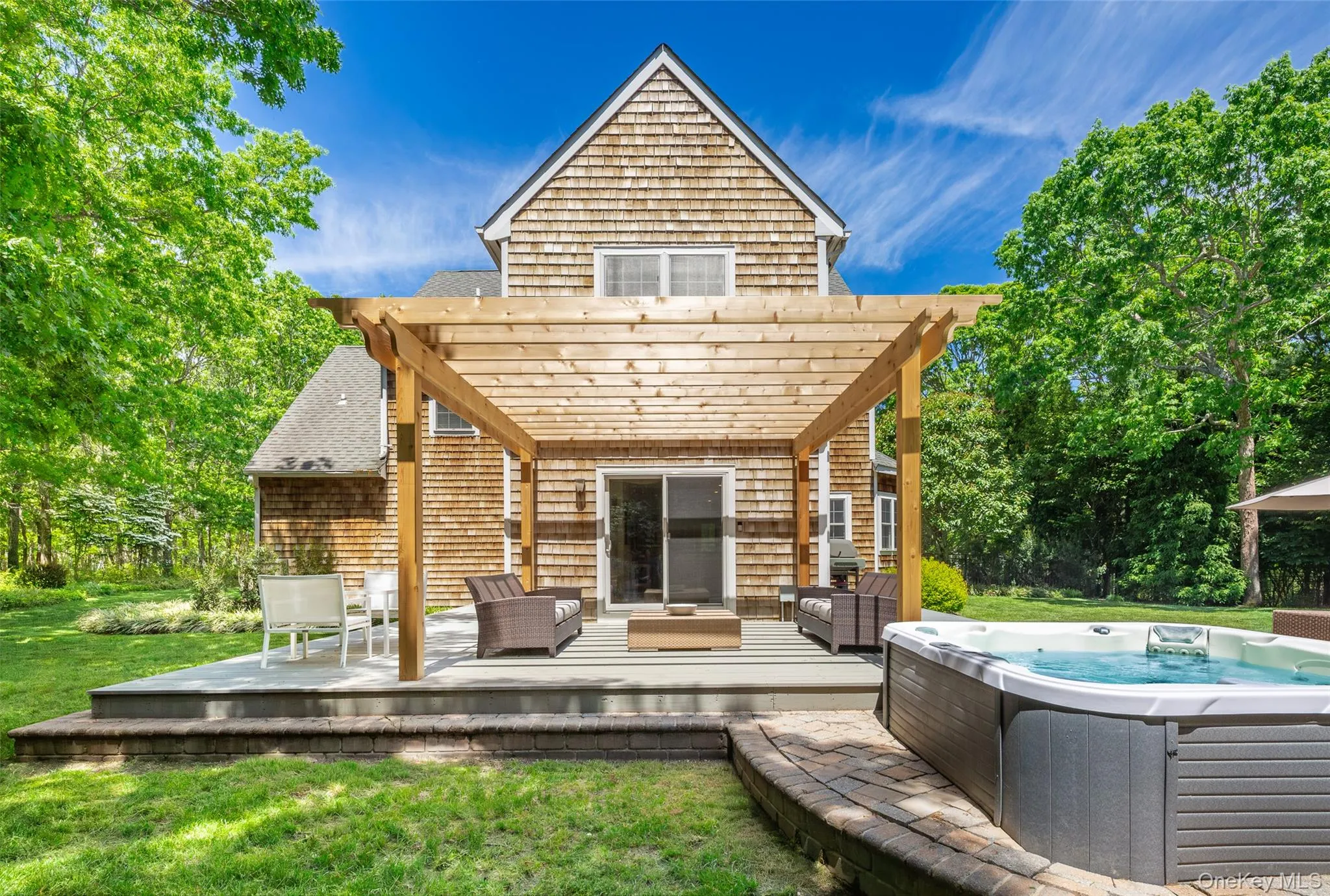 Cedar shake exterior featuring a large elevated deck with a wooden pergola, sliding glass door access, and a built-in hot tub on a brick paver surround Cedar shake exterior featuring a large elevated deck with a wooden pergola, sliding glass door access, and a built-in hot tub on a brick paver surround
