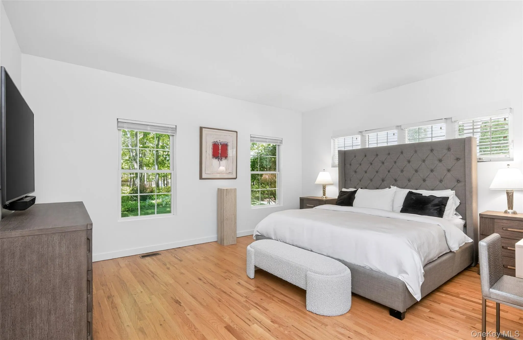 Spacious first floor primary bedroom featuring light wood-finish flooring, white walls, and multiple windows Spacious first floor primary bedroom featuring light wood-finish flooring, white walls, and multiple windows