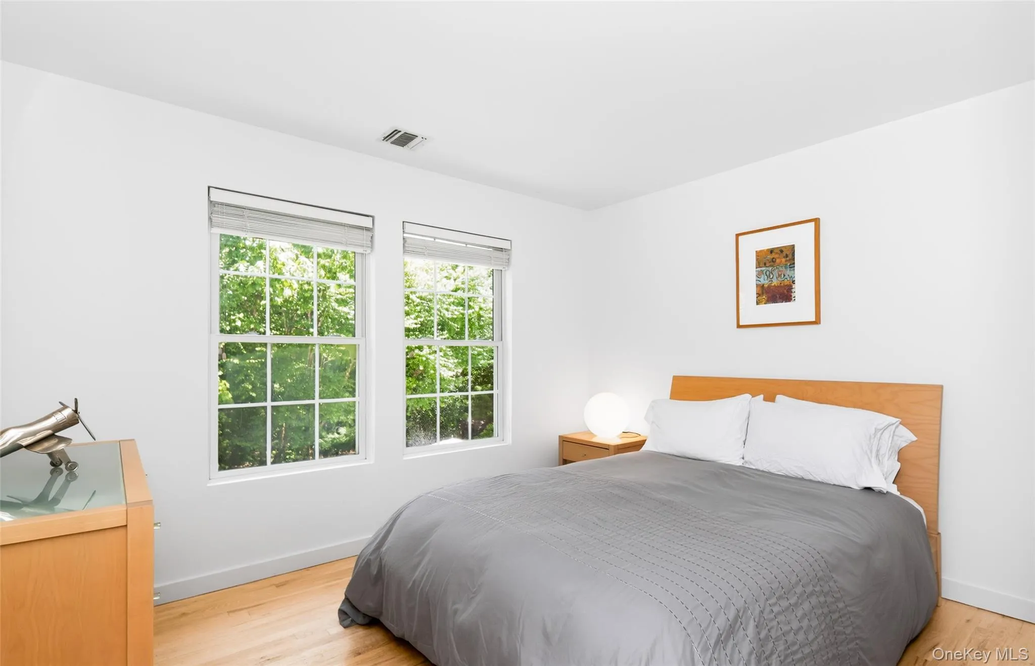 Bright interior space featuring two large windows with natural light, wood-finish flooring, white walls, and a built-in air vent Bright interior space featuring two large windows with natural light, wood-finish flooring, white walls, and a built-in air vent