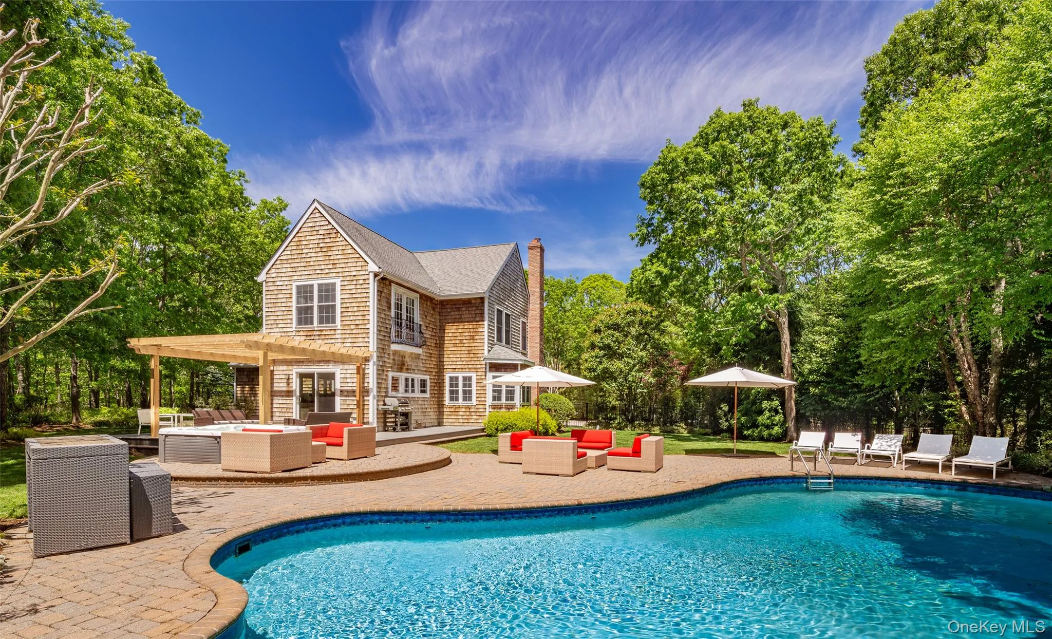 Shingle-style residence featuring an inground swimming pool, paver patio, wood pergola, and mature trees creating a private setting Shingle-style residence featuring an inground swimming pool, paver patio, wood pergola, and mature trees creating a private setting