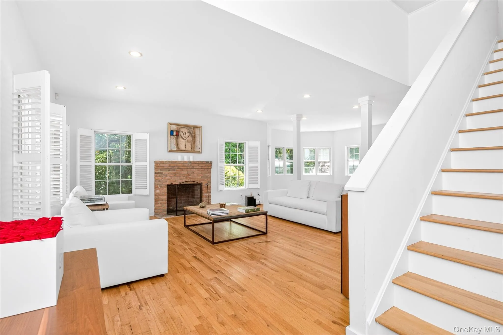 Bright living area featuring wood-finish flooring, a brick fireplace with a metal screen, multiple windows with white plantation shutters, recessed lighting, and a prominent staircase with wood treads Bright living area featuring wood-finish flooring, a brick fireplace with a metal screen, multiple windows with white plantation shutters, recessed lighting, and a prominent staircase with wood treads