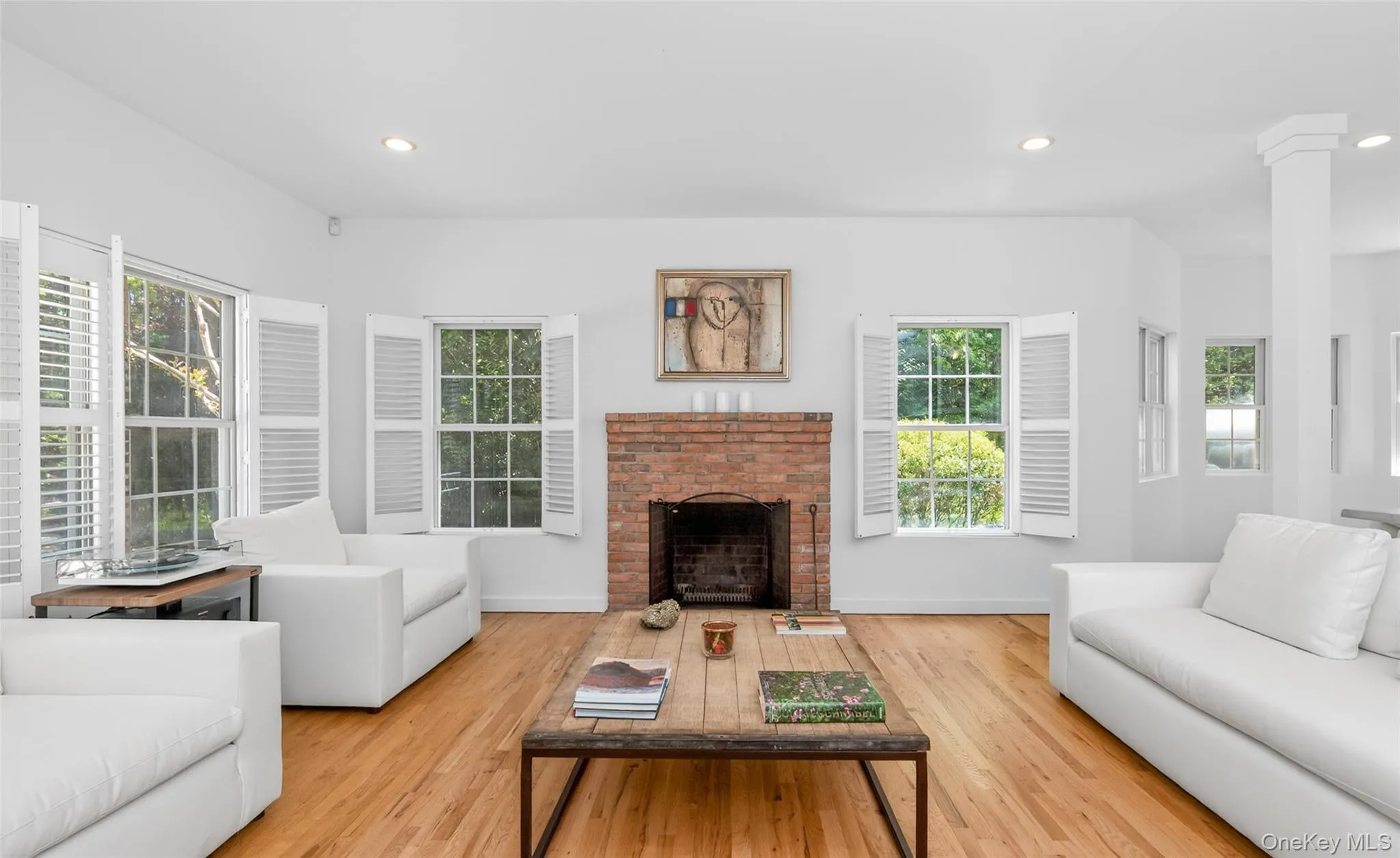Living space featuring a brick fireplace with a wood mantel, hardwood flooring, and multiple windows with interior plantation shutters Living space featuring a brick fireplace with a wood mantel, hardwood flooring, and multiple windows with interior plantation shutters