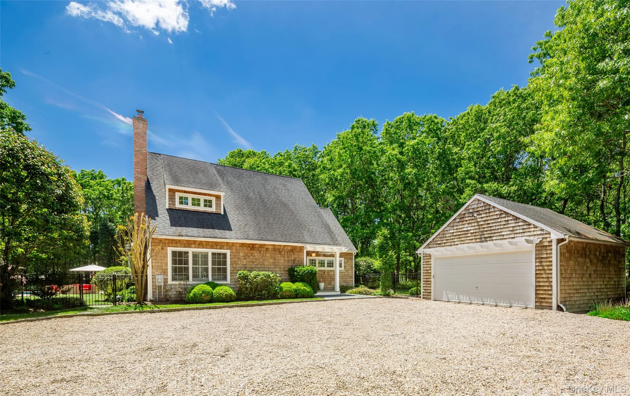 Shingle-style home featuring a prominent brick chimney, dormer window, and a gravel driveway and a 2 car garage Shingle-style home featuring a prominent brick chimney, dormer window, and a gravel driveway and a 2 car garage