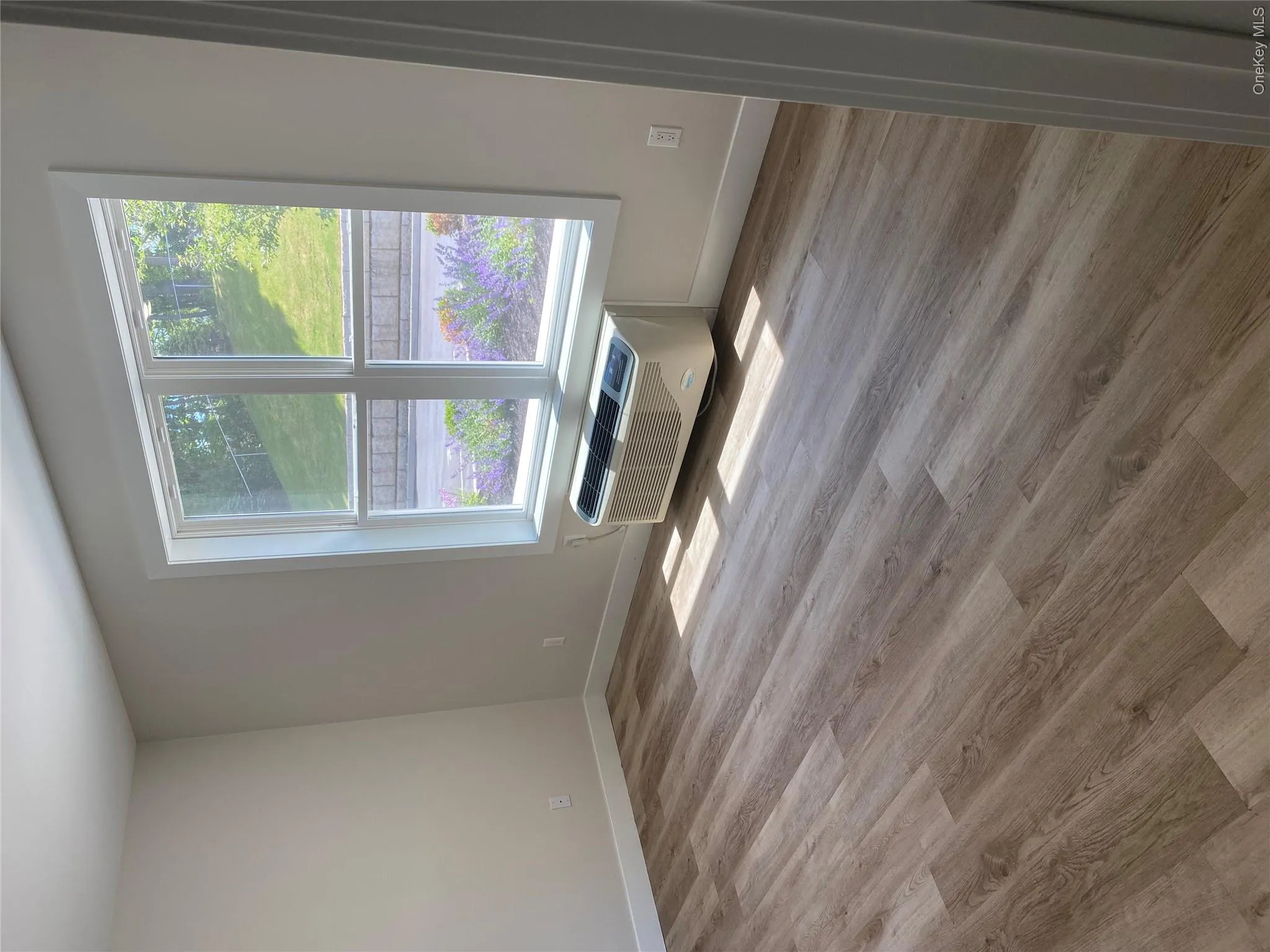 Room featuring wood-finish flooring, a large window with white trim, and a wall-mounted air conditioning unit Room featuring wood-finish flooring, a large window with white trim, and a wall-mounted air conditioning unit