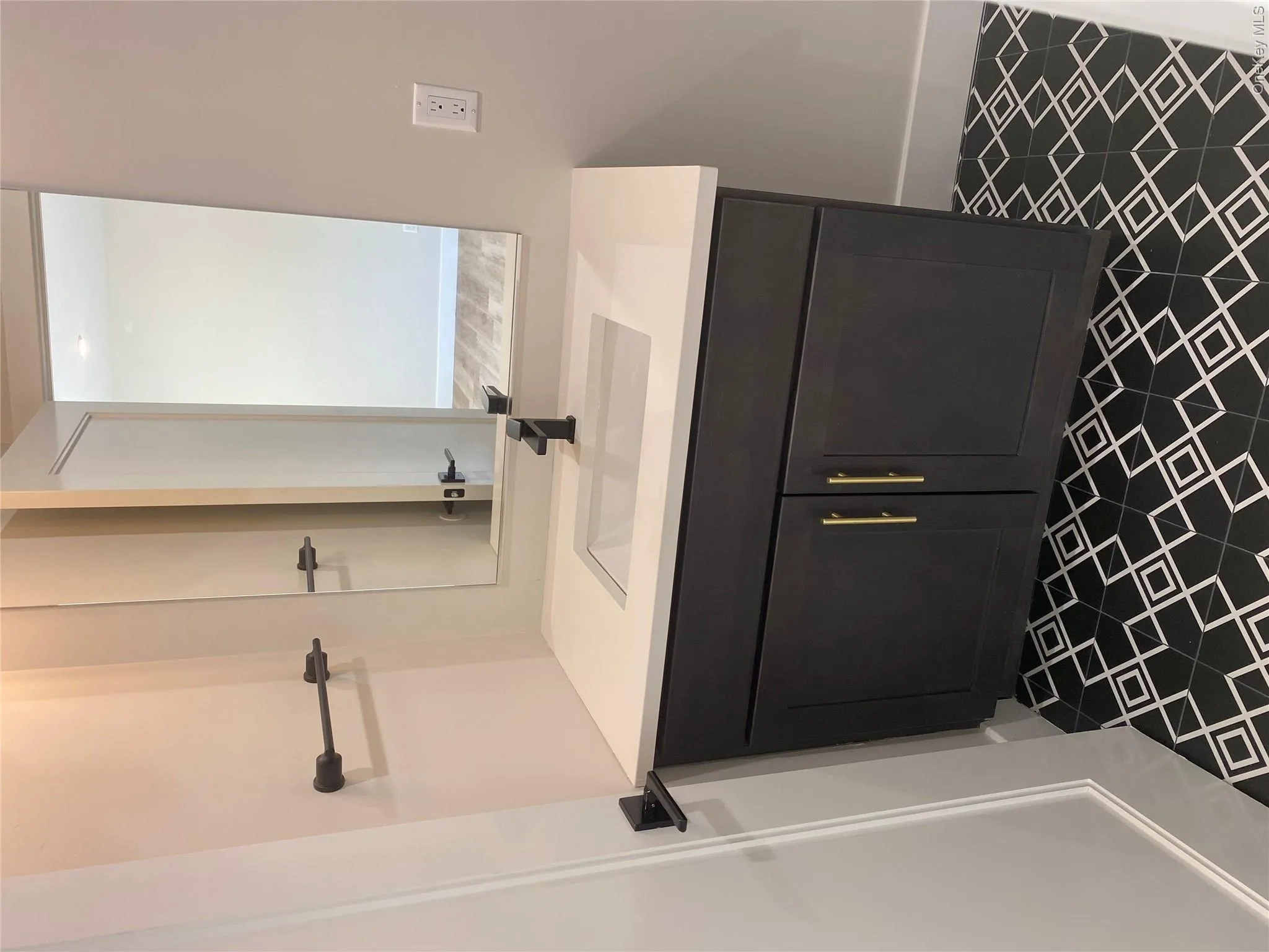 Vanity with a white countertop and integrated sink, black base cabinetry with gold-toned hardware, matte black faucet, and patterned floor tiling Vanity with a white countertop and integrated sink, black base cabinetry with gold-toned hardware, matte black faucet, and patterned floor tiling