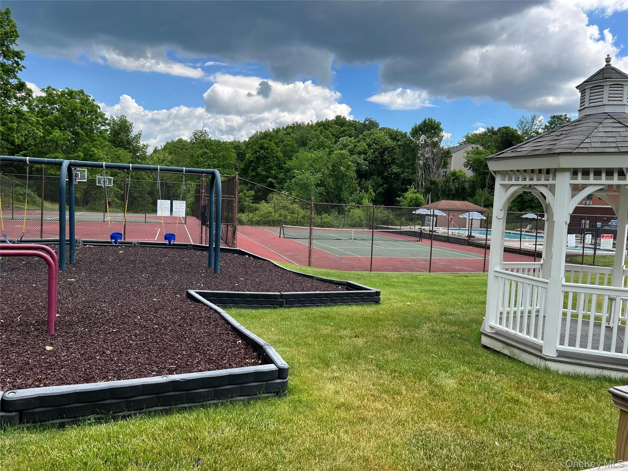 Expansive outdoor recreation area featuring a white wooden gazebo with a shingled roof, a swing set with a wood chip surface, and a fenced-in tennis court Expansive outdoor recreation area featuring a white wooden gazebo with a shingled roof, a swing set with a wood chip surface, and a fenced-in tennis court