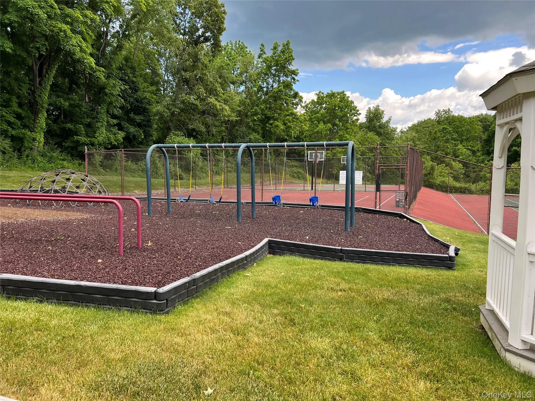Outdoor recreation area featuring a playground with swings, a climbing dome, and a red-surfaced sports court Outdoor recreation area featuring a playground with swings, a climbing dome, and a red-surfaced sports court