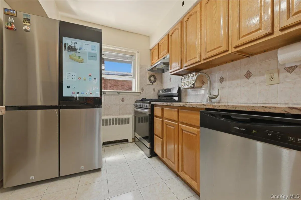 Kitchen featuring stainless steel appliances, wood cabinetry, tile backsplash with accent tiles, light-toned countertops, and tile flooring Kitchen featuring stainless steel appliances, wood cabinetry, tile backsplash with accent tiles, light-toned countertops, and tile flooring