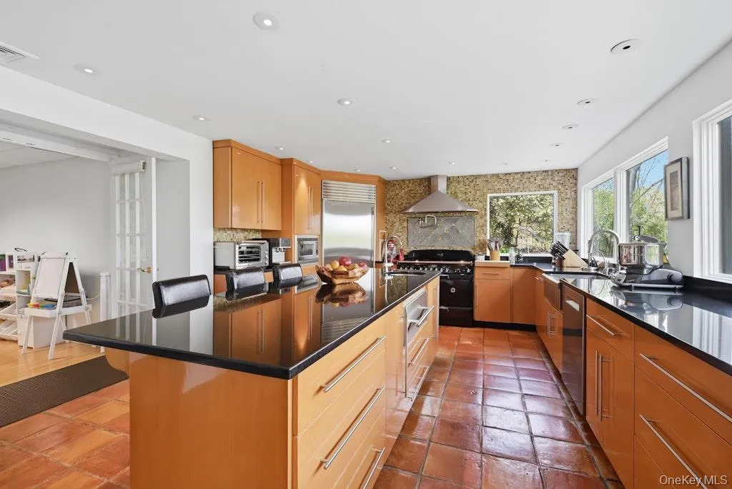 Spacious kitchen featuring an expansive island with a black countertop, natural wood-finish cabinetry, terracotta tile flooring, a large range with a stainless steel vent hood, and multiple windows Spacious kitchen featuring an expansive island with a black countertop, natural wood-finish cabinetry, terracotta tile flooring, a large range with a stainless steel vent hood, and multiple windows