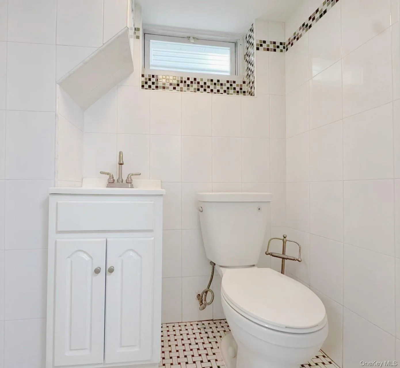 Bathroom featuring white tiled walls, a mosaic tile border, a white vanity with brushed nickel hardware, and black and white basketweave floor tiling Bathroom featuring white tiled walls, a mosaic tile border, a white vanity with brushed nickel hardware, and black and white basketweave floor tiling
