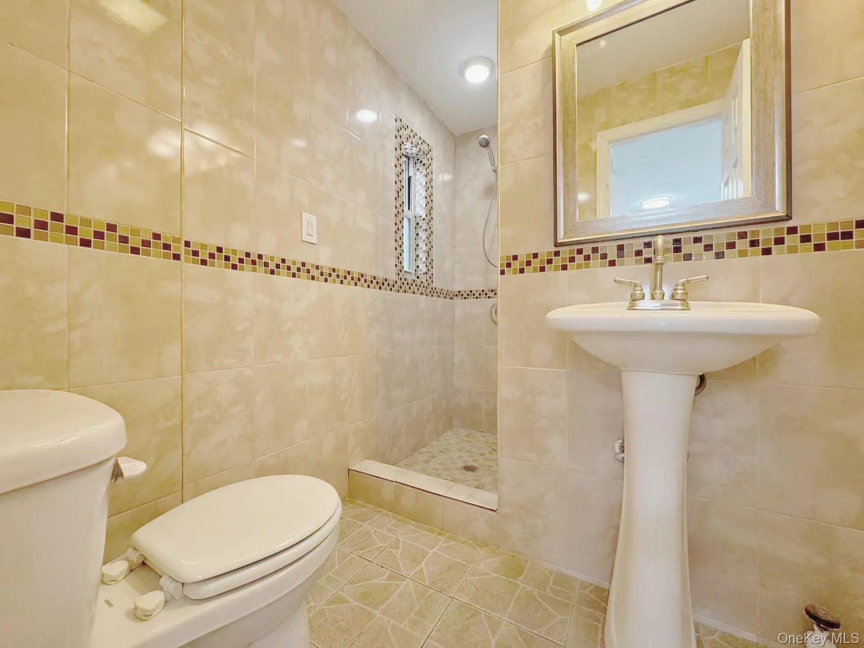 Bathroom featuring a pedestal sink, walk-in shower with mosaic tile accents, and tiled walls with a decorative border Bathroom featuring a pedestal sink, walk-in shower with mosaic tile accents, and tiled walls with a decorative border