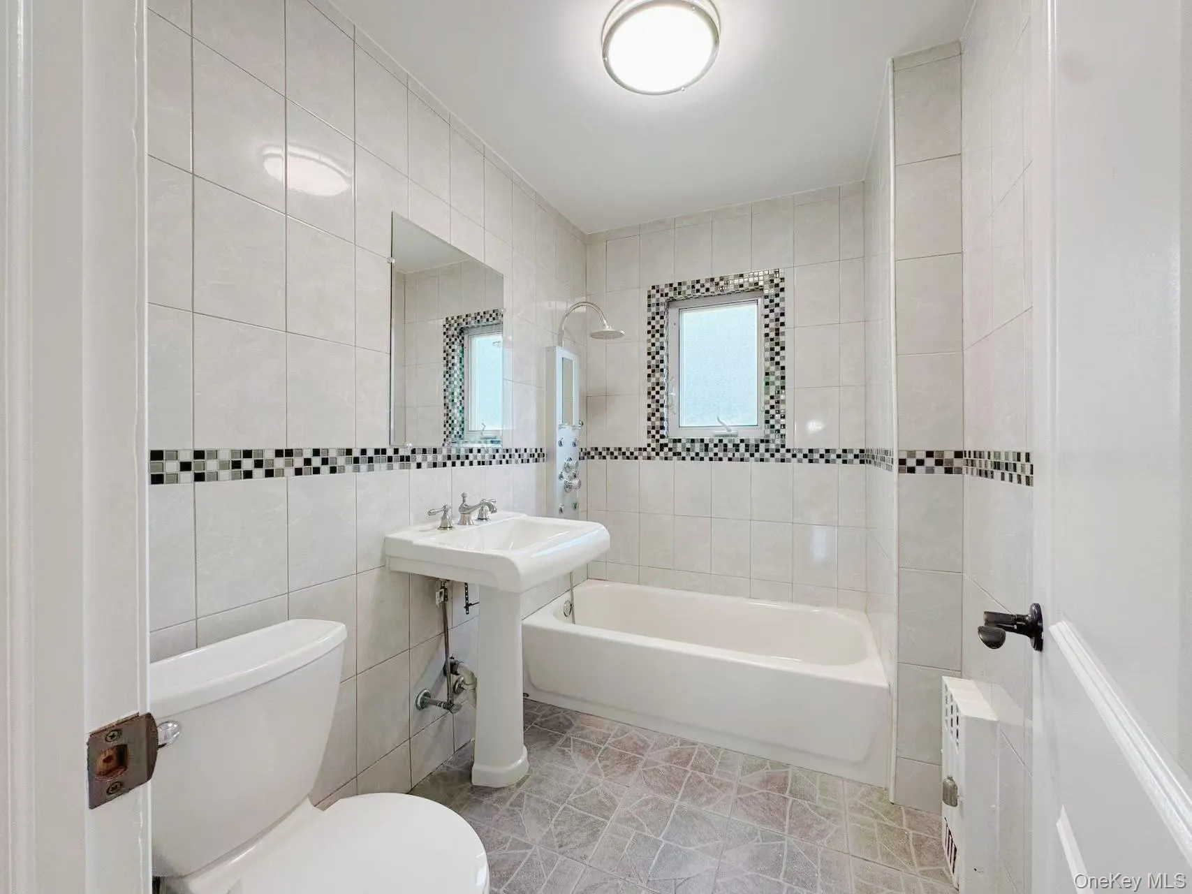 Bathroom featuring full wall tiling with a decorative mosaic band, pedestal sink with chrome faucet, bathtub with shower fixture, and a window with mosaic tile surround Bathroom featuring full wall tiling with a decorative mosaic band, pedestal sink with chrome faucet, bathtub with shower fixture, and a window with mosaic tile surround