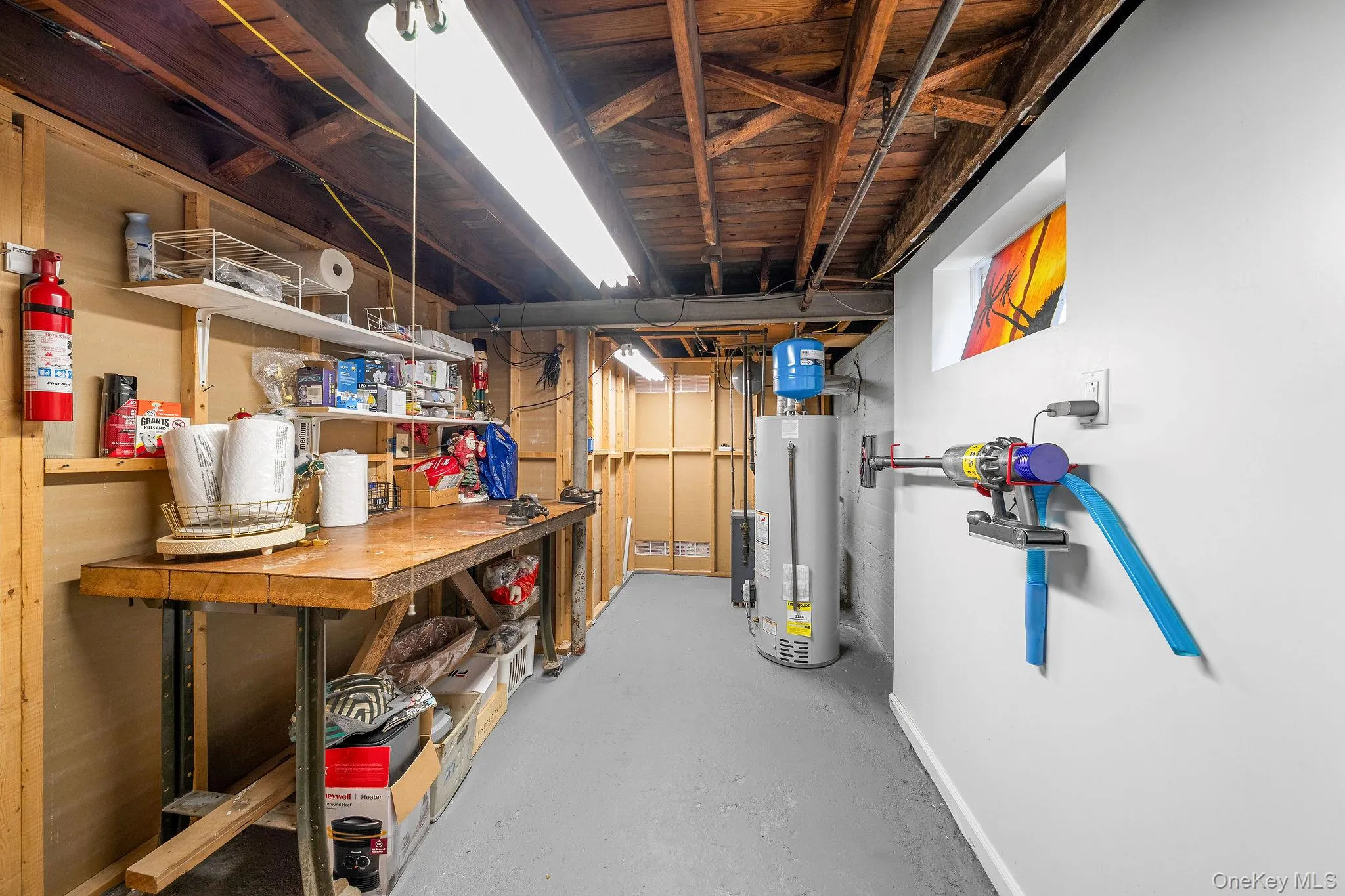 Utility space featuring exposed wood ceiling joists, durable gray flooring, a built-in workbench, shelving, and well maintained water heater. Utility space featuring exposed wood ceiling joists, durable gray flooring, a built-in workbench, shelving, and well maintained water heater.