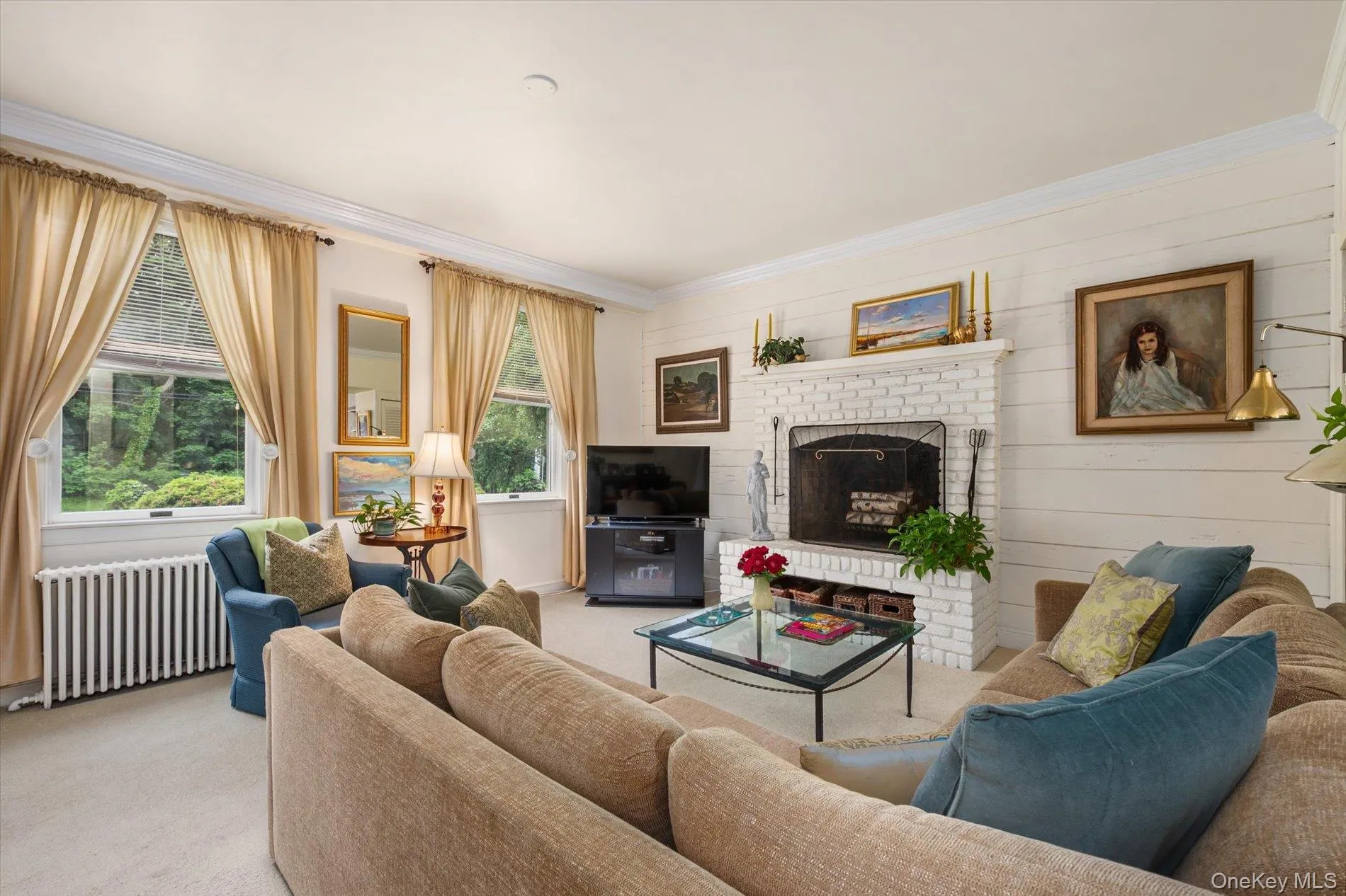 Living area featuring a painted brick fireplace with a metal grate, white shiplap accent wall, crown molding, and multiple windows Living area featuring a painted brick fireplace with a metal grate, white shiplap accent wall, crown molding, and multiple windows