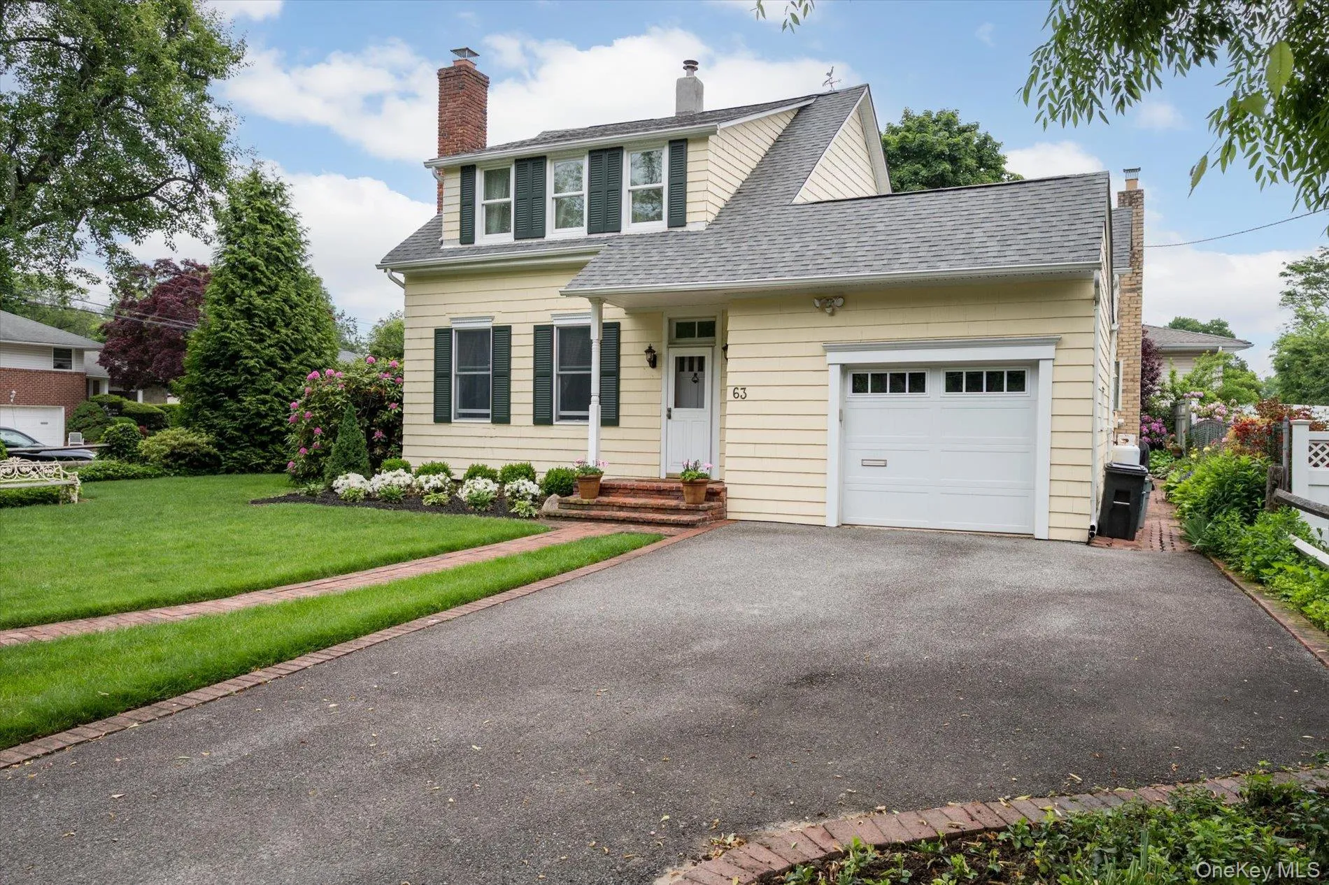 Cream-colored siding home featuring a front-facing garage with a paneled door, dark green window shutters, a red brick chimney, and a landscaped front yard with a paved driveway Cream-colored siding home featuring a front-facing garage with a paneled door, dark green window shutters, a red brick chimney, and a landscaped front yard with a paved driveway